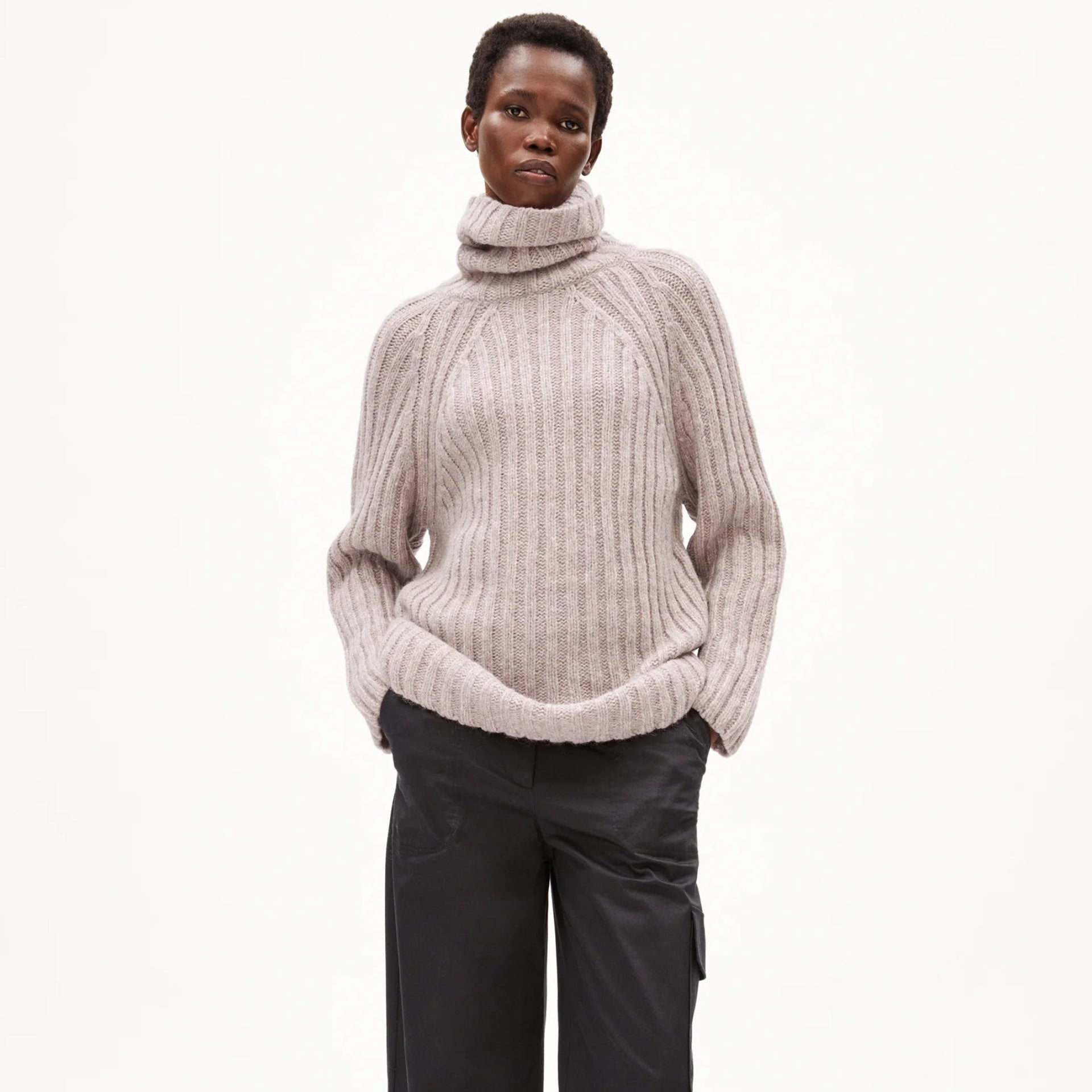 Eine Person steht mit den Händen in den Taschen, trägt einen dicken hellbeigen Rollkragenpullover, eine weite dunkle Hose und gestreifte Turnschuhe vor einem schlichten weißen Hintergrund.