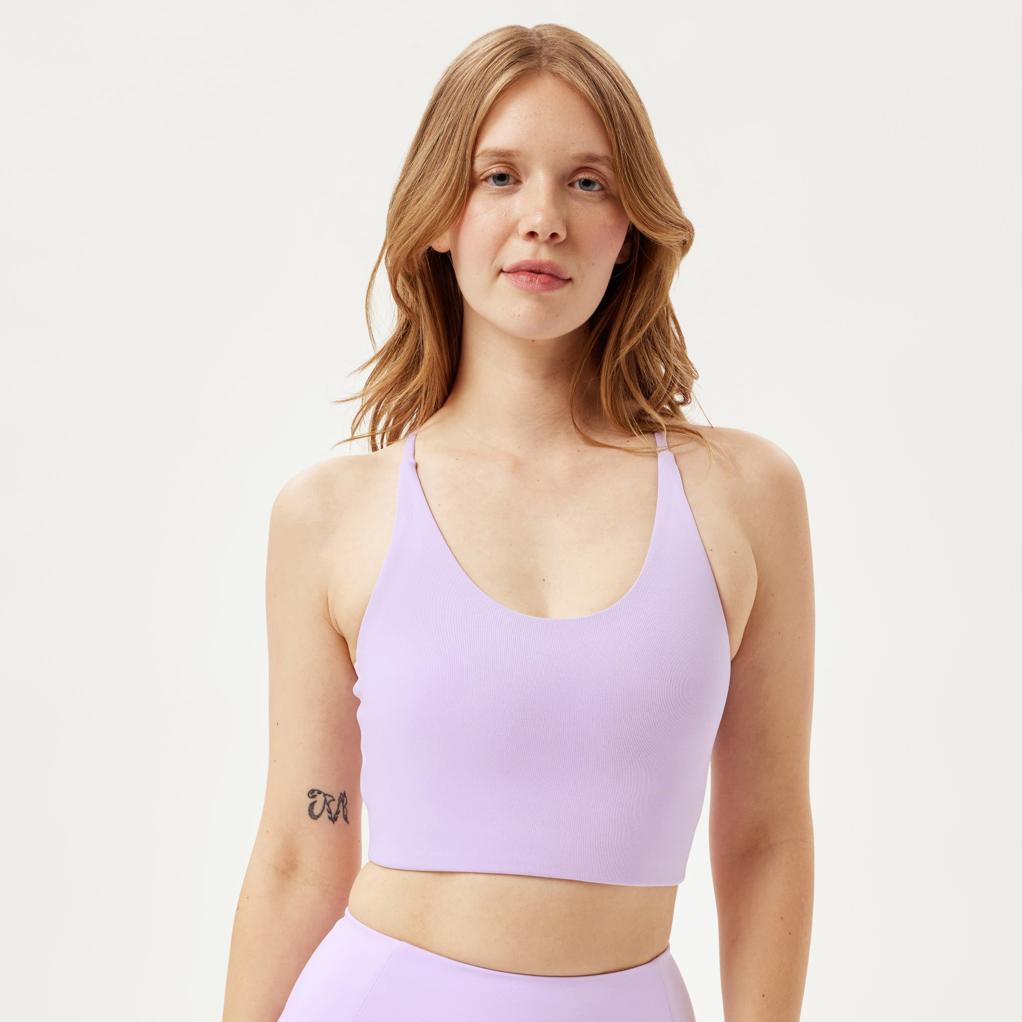 Eine Frau mit hellbraunem Haar, die einen lavendelfarbenen Sport-BH und dazu passende Leggings trägt, steht mit dem Gesicht nach vorne vor einem schlichten weißen Hintergrund, auf dem eine kleine Tätowierung auf ihrem rechten Brustkorb zu sehen ist.