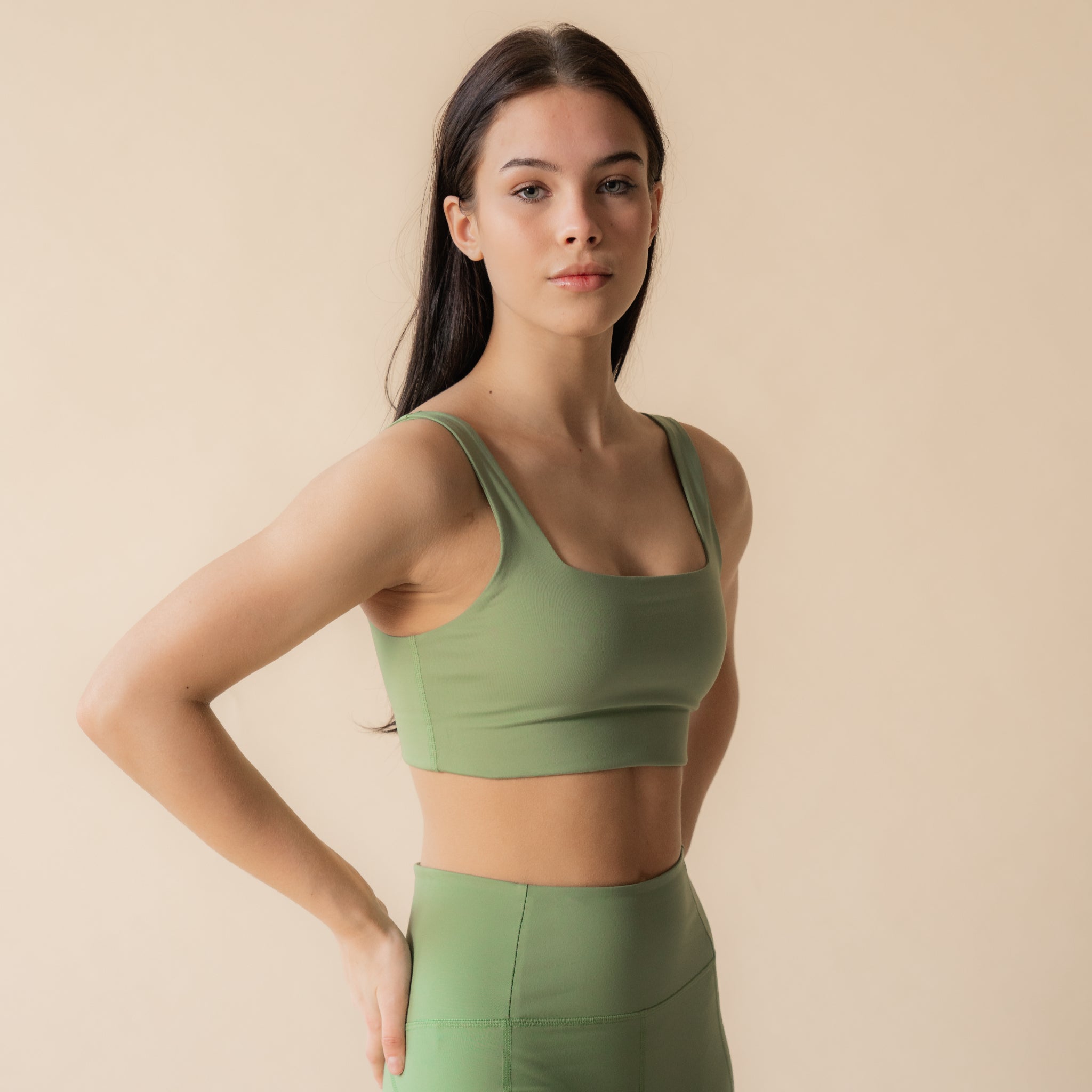 Eine Frau mit langen dunklen Haaren, die einen grünen Sport-BH und dazu passende Leggings trägt, steht vor einem beigen Hintergrund und blickt selbstbewusst in die Kamera, wobei sie eine Hand auf ihre Hüfte stützt.