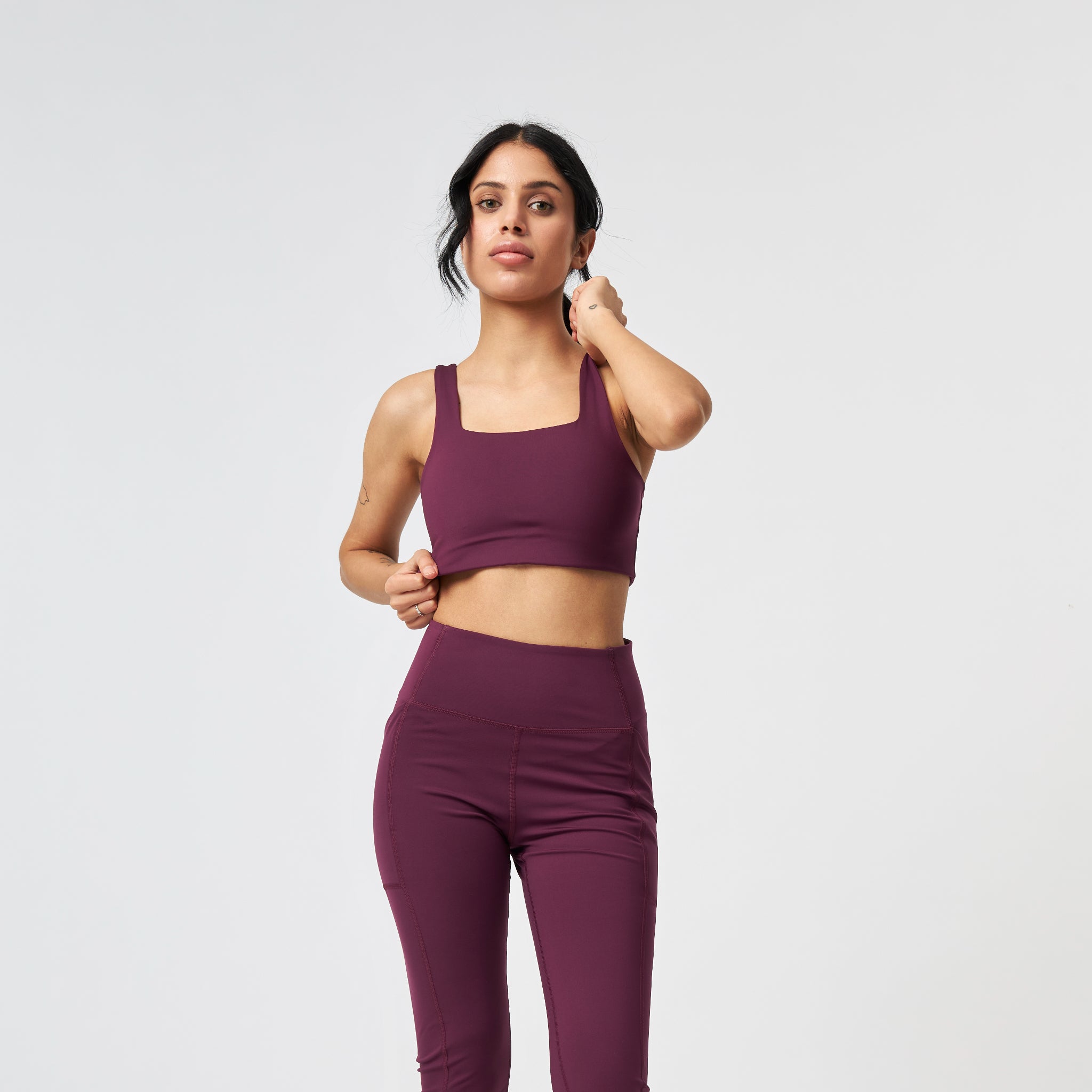 Eine Frau, die einen passenden kastanienbraunen Sport-BH und hoch taillierte Leggings trägt, steht vor einem schlichten weißen Hintergrund und blickt nach vorne, wobei sie mit einer Hand ihr Haar berührt.