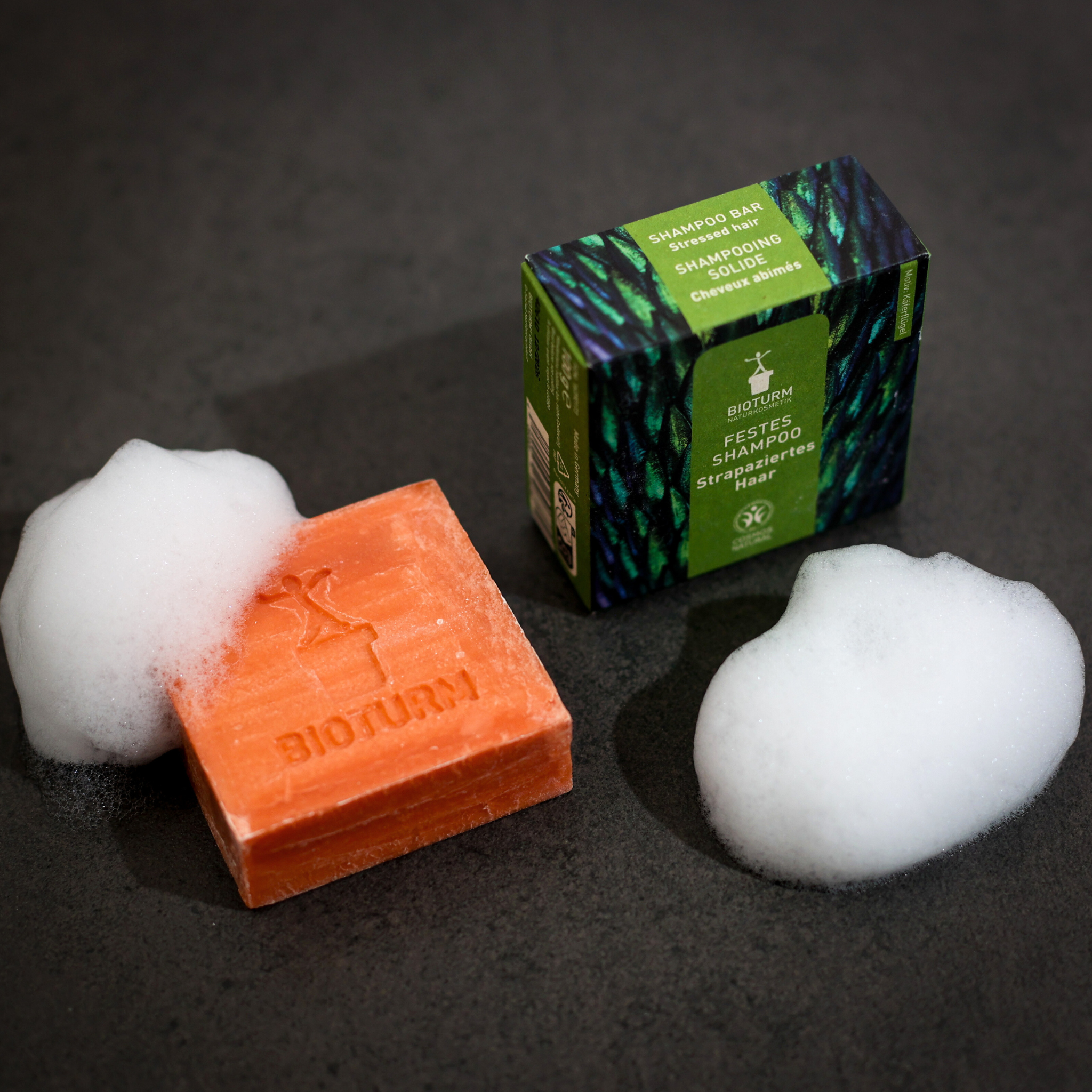 Eine orangefarbene feste Shampooseife mit Schaum neben einer grün-schwarzen Shampoo-Verpackung auf grauem Untergrund.