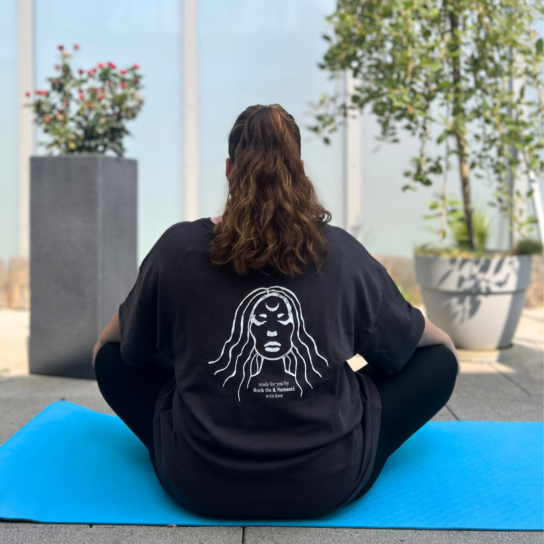 Eine Person sitzt auf einer blauen Yogamatte mit dem Rücken zur Kamera, umgeben von Pflanzen in Töpfen.