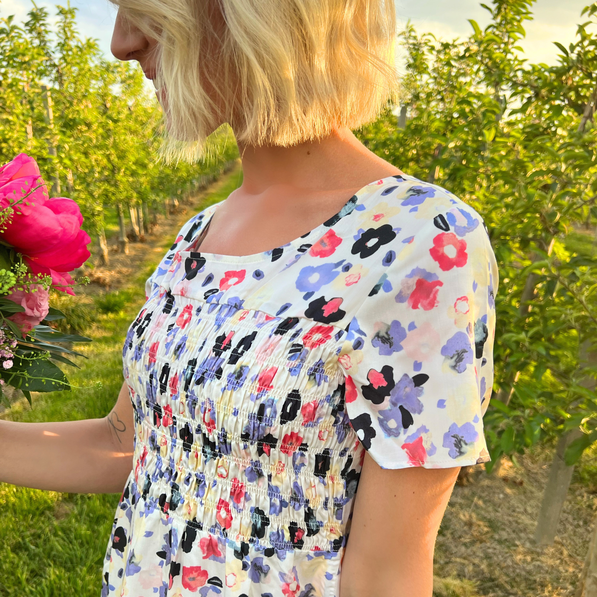 Eine Frau mit blonden Haaren in einem Blumenfeld hält einen Blumenstrauß und trägt ein geblümtes Kleid.