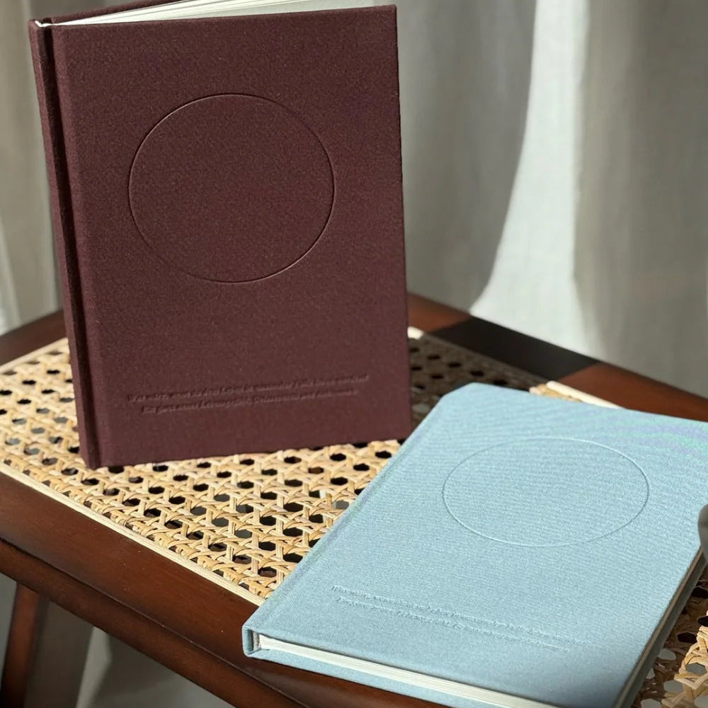 Zwei strukturierte Hardcover-Bücher, ein weinrotes aufrecht stehend und ein hellblaues liegend, sitzen auf einem Holzstuhl mit geflochtenem Rattansitz, der in natürliches Sonnenlicht getaucht ist.