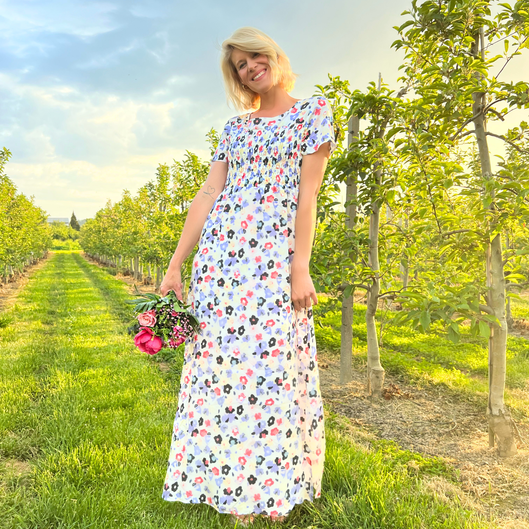 Eine Person in einem geblümten Kleid steht lächelnd in einem Obstgarten und hält einen Blumenstrauß in der Hand.