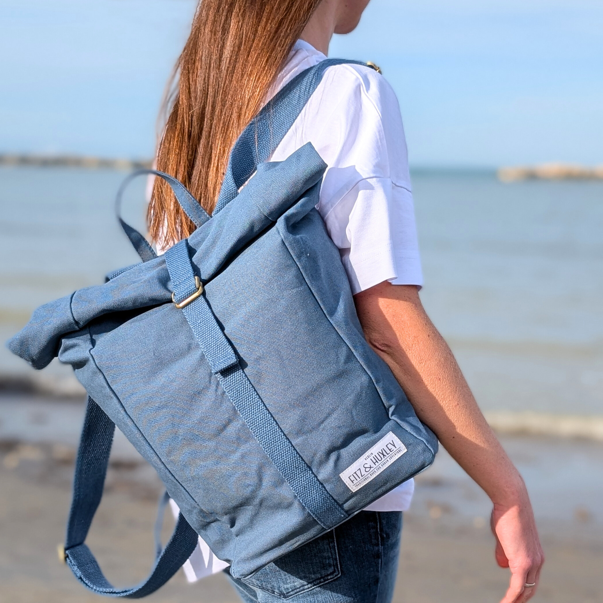 Frau mit blauem Rucksack geht am Strand entlang, im Hintergrund das Meer.