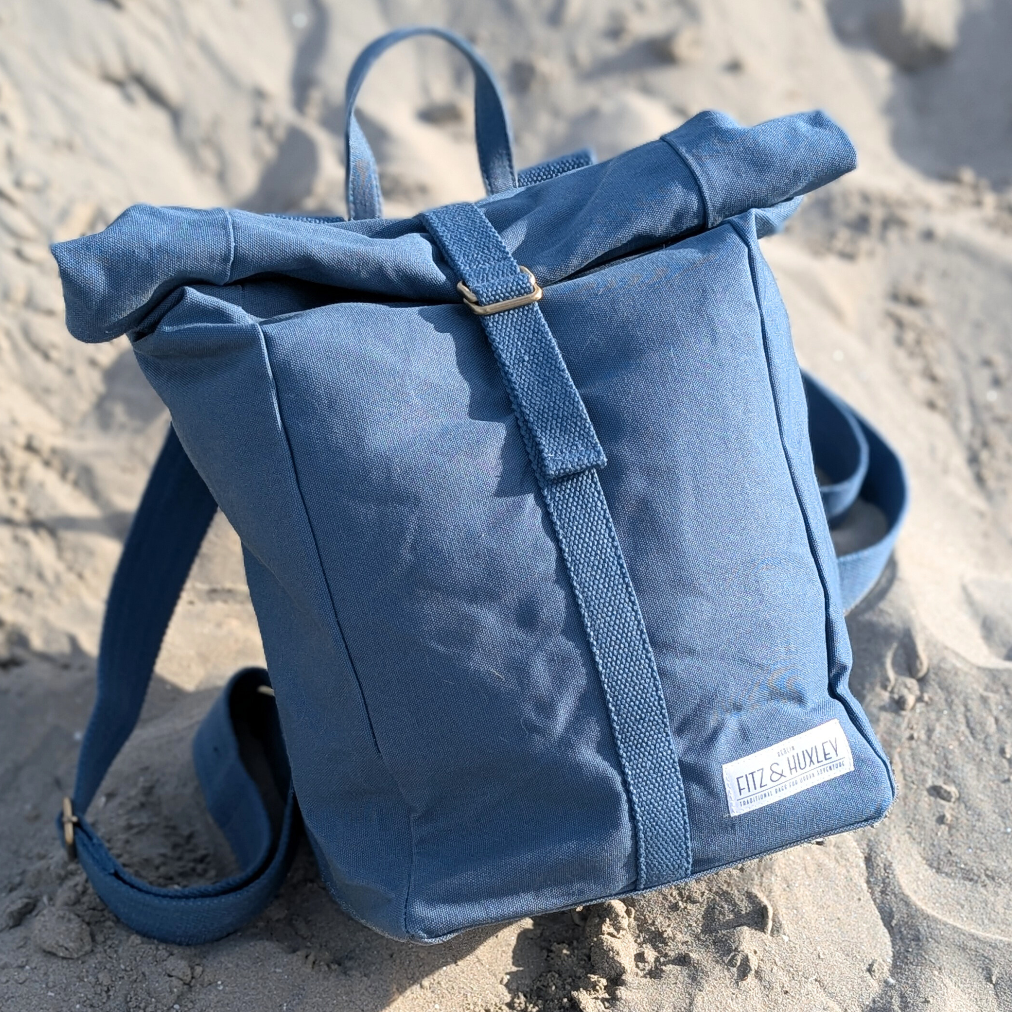Blauer Rucksack auf sandigem Boden, mit markantem Roll-Top-Verschluss und Logo auf der Vorderseite.