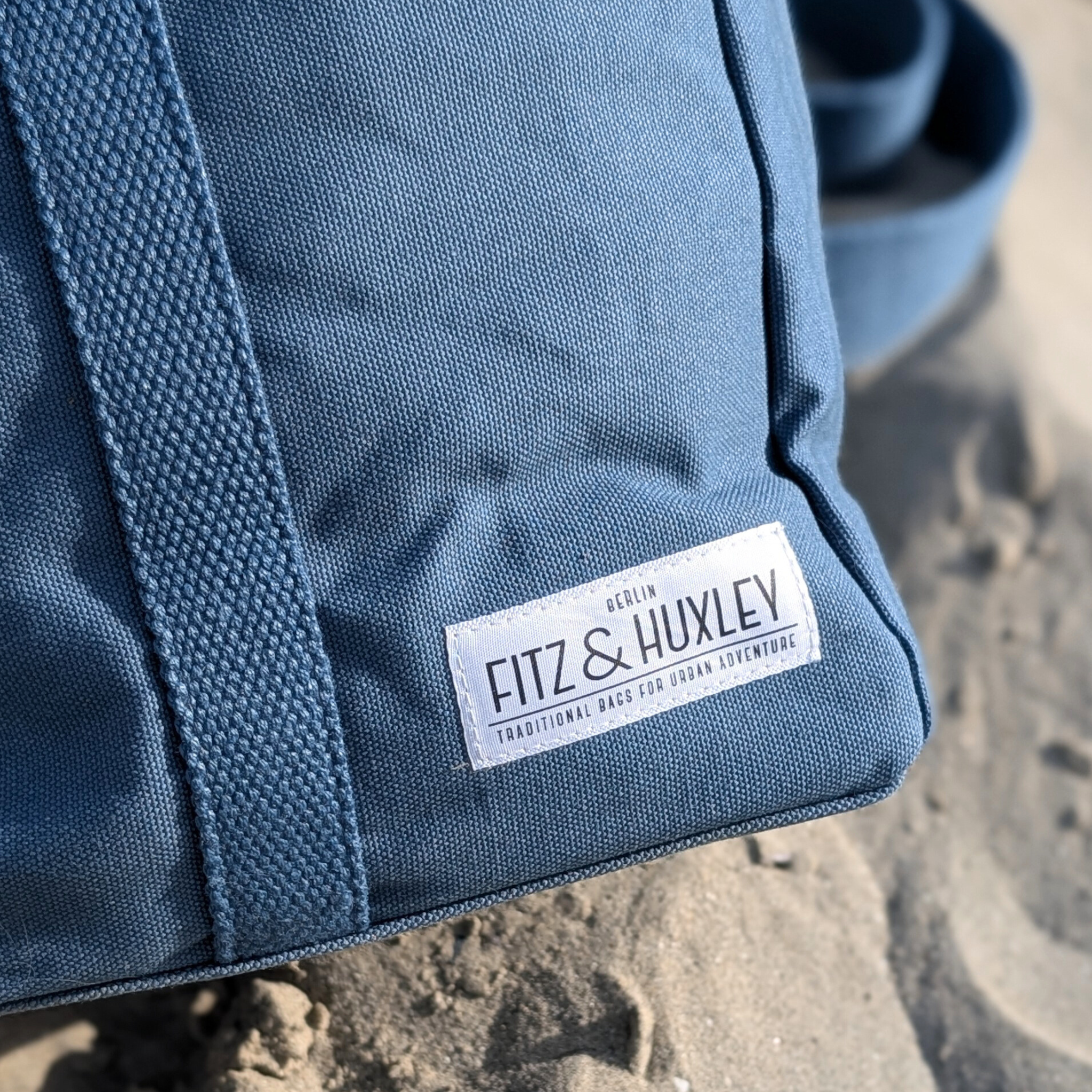 Blauer Stoffrucksack auf Sand mit einem Etikett der Marke Fitz & Huxley aus Berlin.