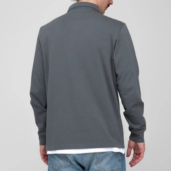 Eine Person, die ein langärmeliges graues Hemd und eine blaue Jeans trägt, steht mit leicht zur Seite gedrehtem Kopf vor einem schlichten, hellen Hintergrund und ist von der Kamera abgewandt.