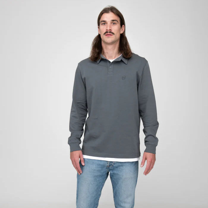 Ein Mann mit langen braunen Haaren und Schnurrbart trägt ein graues langärmeliges Poloshirt über einem weißen Unterhemd und hellblaue Jeans vor einem schlichten hellgrauen Hintergrund.