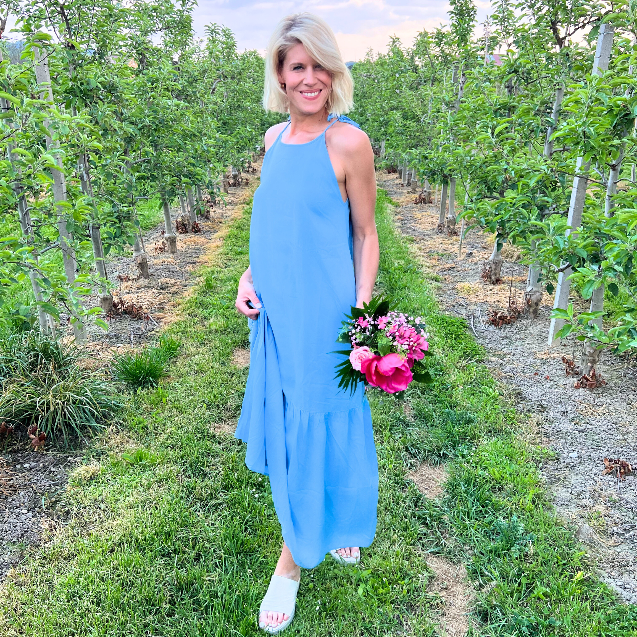 Frau im blauen Kleid steht mit Blumenstrauß in einem Obstgarten und lächelt.