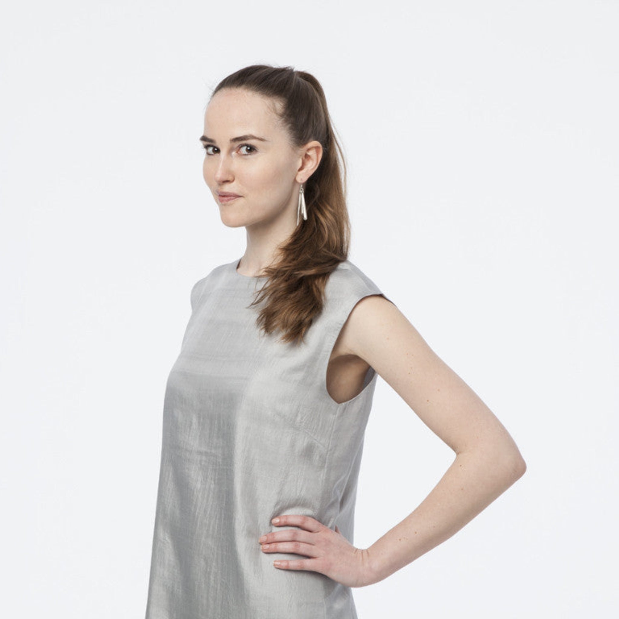 Frau in einem ärmellosen, grauen Kleid vor weißem Hintergrund, die zur Seite schaut und lächelt.