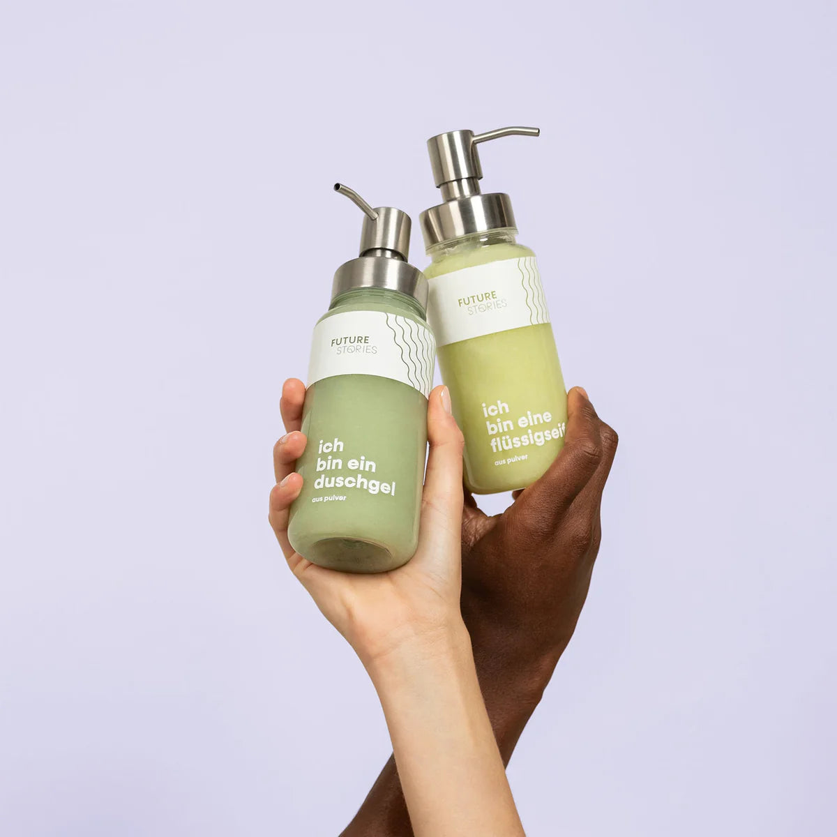 Zwei Hände mit unterschiedlichen Hauttönen halten grünliche Flüssigseifenflaschen mit Pumpspender vor einem hellvioletten Hintergrund hoch. Die Flaschen sind auf Deutsch mit "Future Stories" beschriftet.