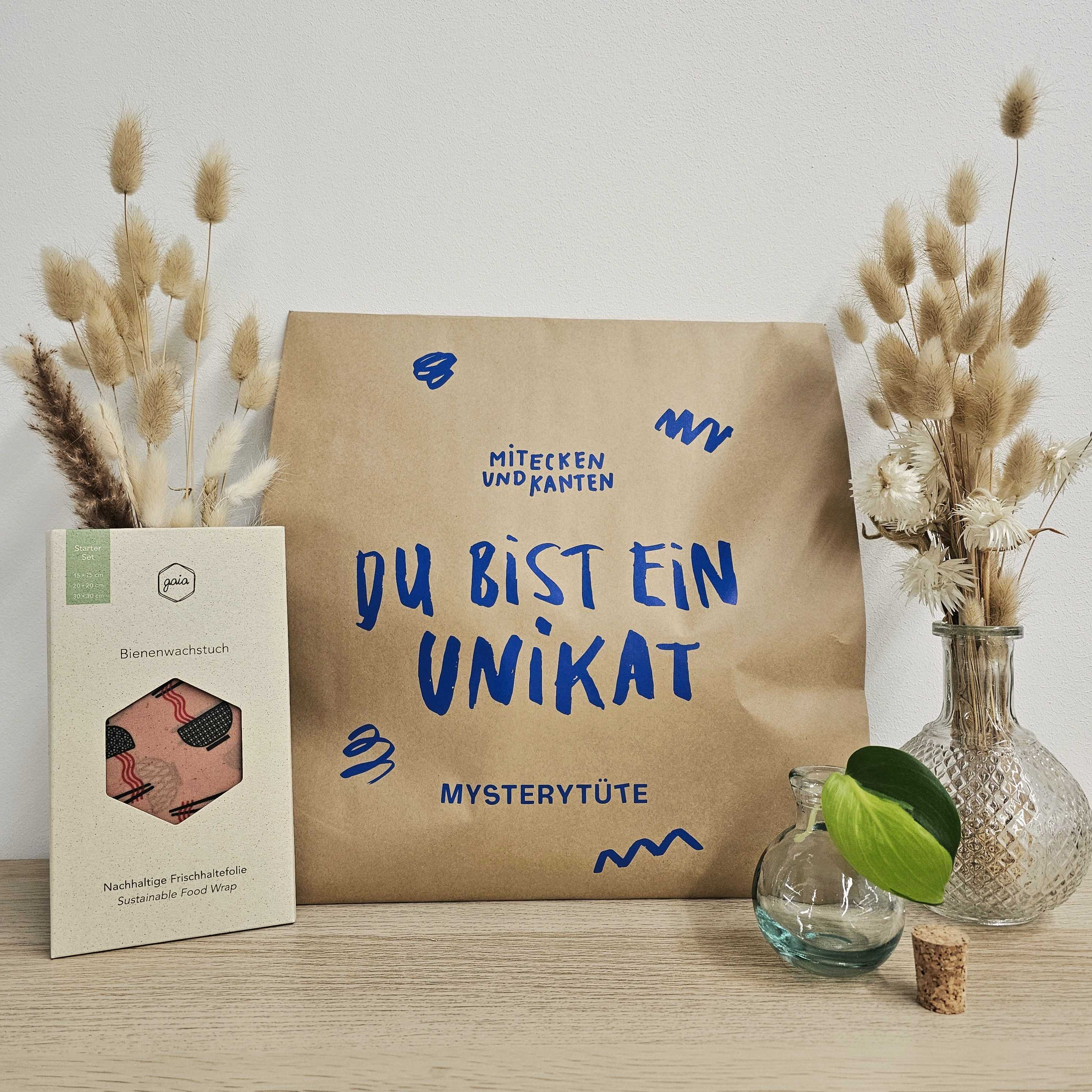 Eine braune Papiertüte mit blauer Aufschrift Du bist ein Unikat liegt auf einer Holzfläche zwischen getrockneten Blumenarrangements in Glasvasen. Links daneben liegt eine Bienenwachsverpackung mit Bienenmotiv und deutschem Text.