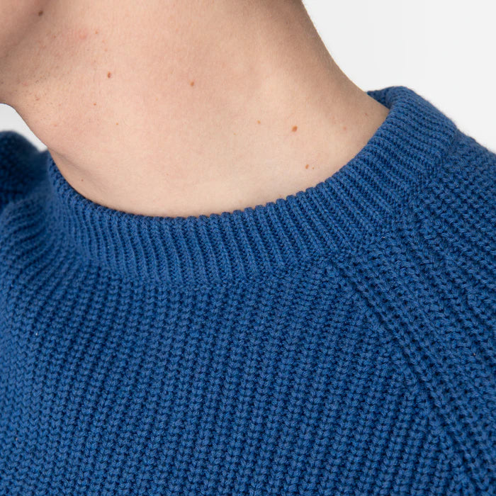 Nahaufnahme von Hals und Schulter einer Person, die einen blauen Strickpullover mit Struktur trägt. Die Haut ist hell und es sind einige kleine Leberflecke zu sehen. Das Bild konzentriert sich auf den gerippten Kragen und das Muster des Pullovers.