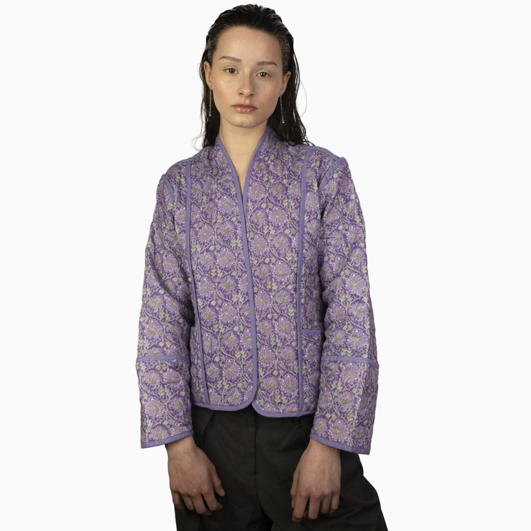Eine Person mit nassem, dunklem Haar trägt eine lavendelfarbene Steppjacke mit Blumenmuster und hellem Besatz und steht mit neutralem Gesichtsausdruck vor einem schlichten hellen Hintergrund.
