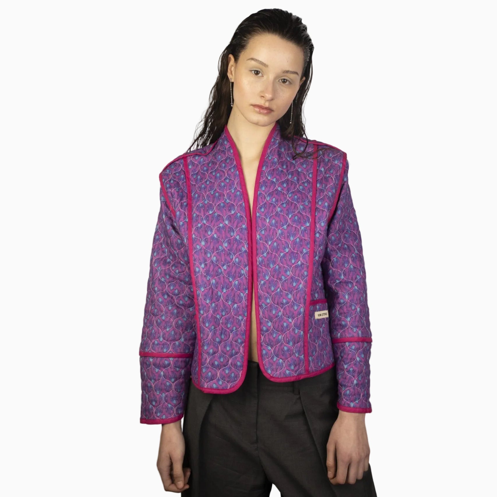 Eine Person mit nassen, zurückgekämmten Haaren trägt eine lila-rosa Steppjacke mit Blumenmuster und eine dunkle Hose vor einem schlichten hellen Hintergrund.