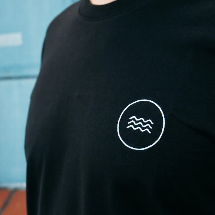 Eine Person trägt ein schwarzes T-Shirt mit einem kleinen, weißen, kreisförmigen Emblem mit drei Wellenlinien im linken Brustbereich. Der Hintergrund ist unscharf.