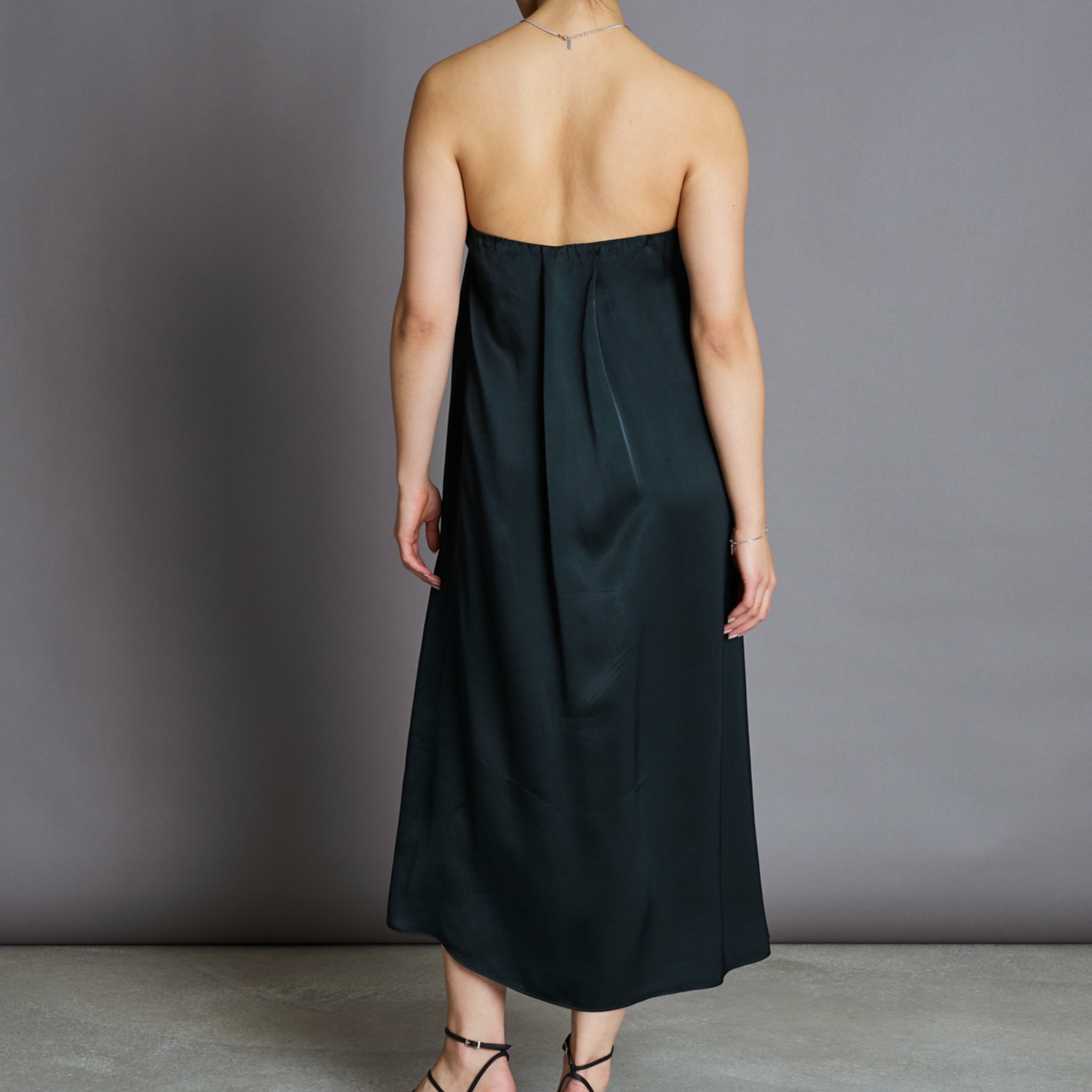 Person in schulterfreiem, langem, dunkelgrünem Kleid von hinten, steht vor grauem Hintergrund.