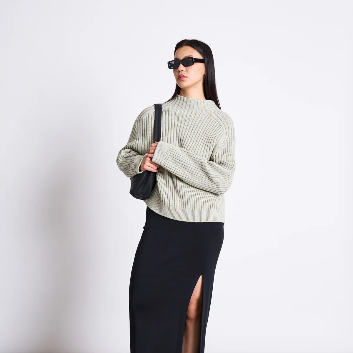Eine Frau, die eine Sonnenbrille, einen hellen gerippten Pullover und einen langen schwarzen Rock mit oberschenkelhohem Schlitz trägt, steht vor einem unifarbenen Hintergrund, hält eine schwarze Umhängetasche und trägt schwarze Slide-Sandalen.
