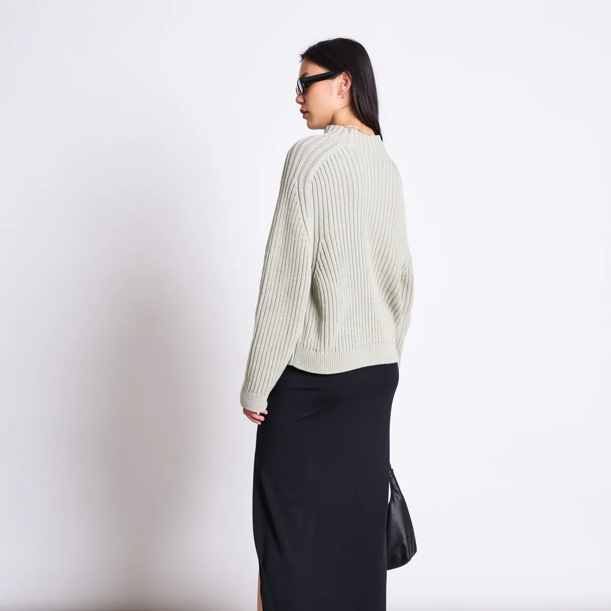 Eine Person, die eine Sonnenbrille, einen hellen gerippten Pullover, einen langen schwarzen Rock mit Seitenschlitz und schwarze Plateausandalen trägt und eine schwarze Tasche in der Hand hält, steht in einem minimalistischen Raum und wendet sich ab.