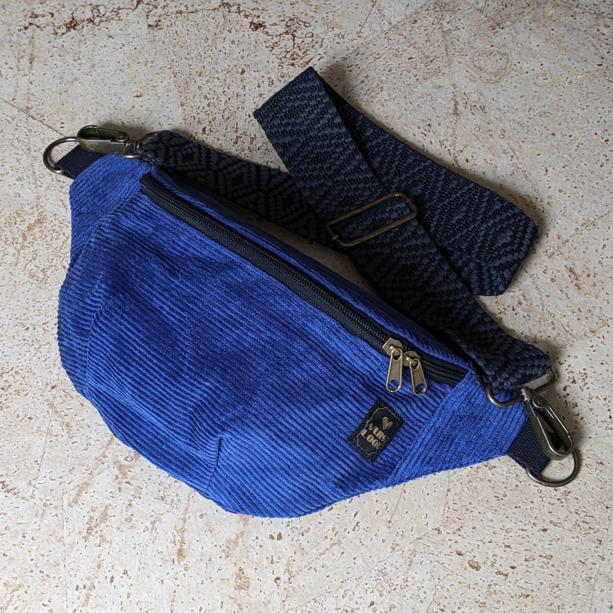 Eine blaue Gürteltasche aus Cord mit einem schwarz gemusterten, verstellbaren Riemen und Metallverschlüssen, die auf einer leicht strukturierten Oberfläche ruht. Die Tasche hat zwei Reißverschlusszüge und ein kleines schwarzes Etikett.