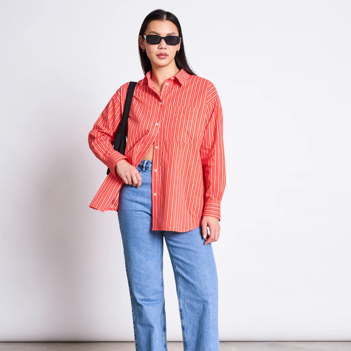 Eine Frau mit einer schwarzen Sonnenbrille, einem rot-weiß gestreiften Button-up-Hemd, hellblauen Jeans und einer schwarzen Umhängetasche steht vor einem schlichten Hintergrund und blickt selbstbewusst in die Kamera.