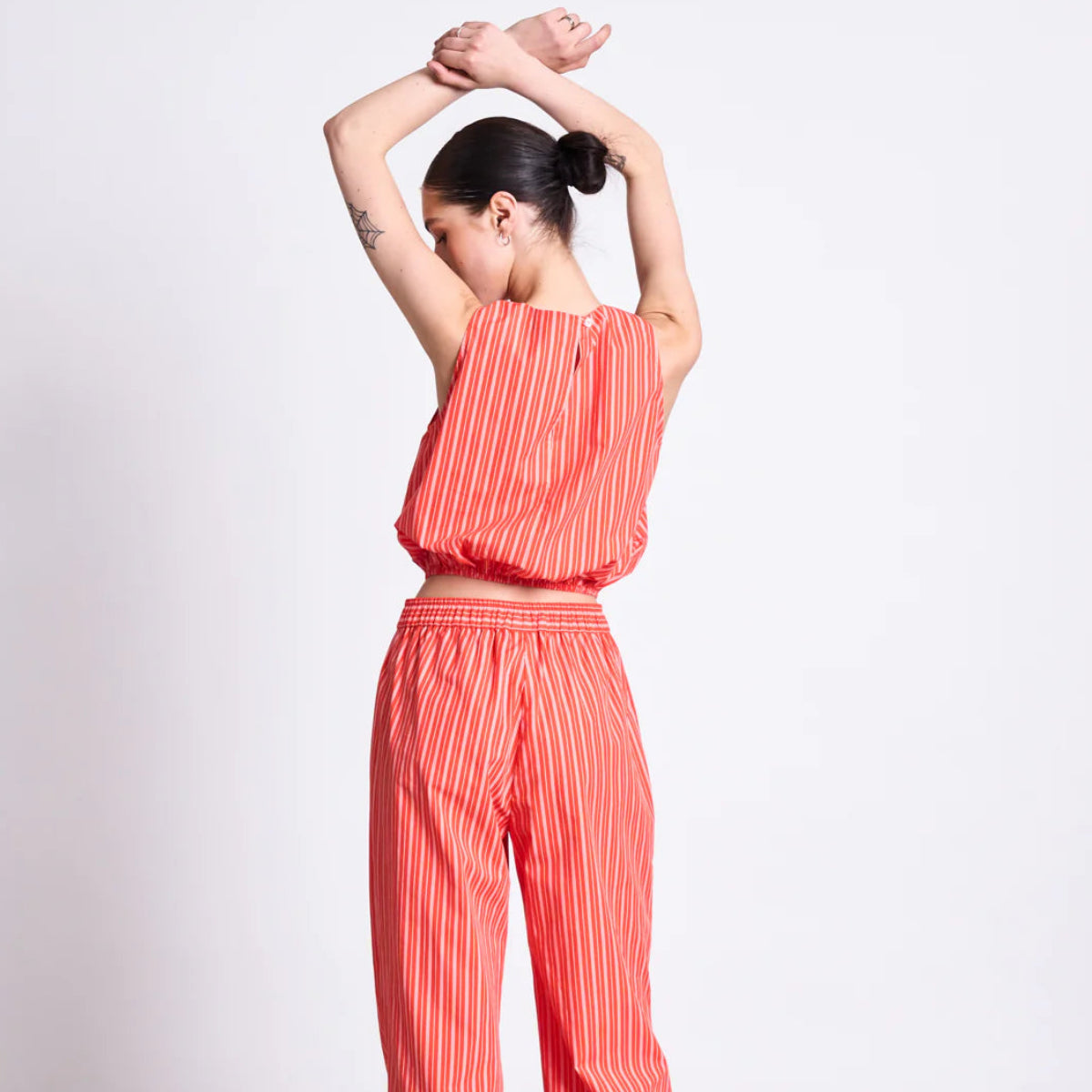 Eine Person steht mit dem Rücken zur Kamera, die Arme erhoben und über dem Kopf verschränkt. Sie trägt ein rotes ärmelloses Oberteil und eine dazu passende Hose mit weiten Beinen und weißen vertikalen Streifen, die Haare sind zu einem tiefen Dutt gebunden.