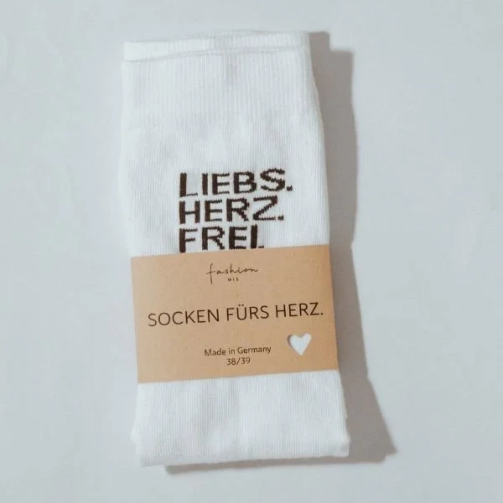 Ein Paar weiße Socken, gefaltet mit einem braunen Papieretikett mit der Aufschrift SOCKEN FÜRS HERZ. Hergestellt in Deutschland 38/39. Die Socken haben einen schwarzen Text mit der Aufschrift LIEBS. HERZ. FREI. aufgedruckt.