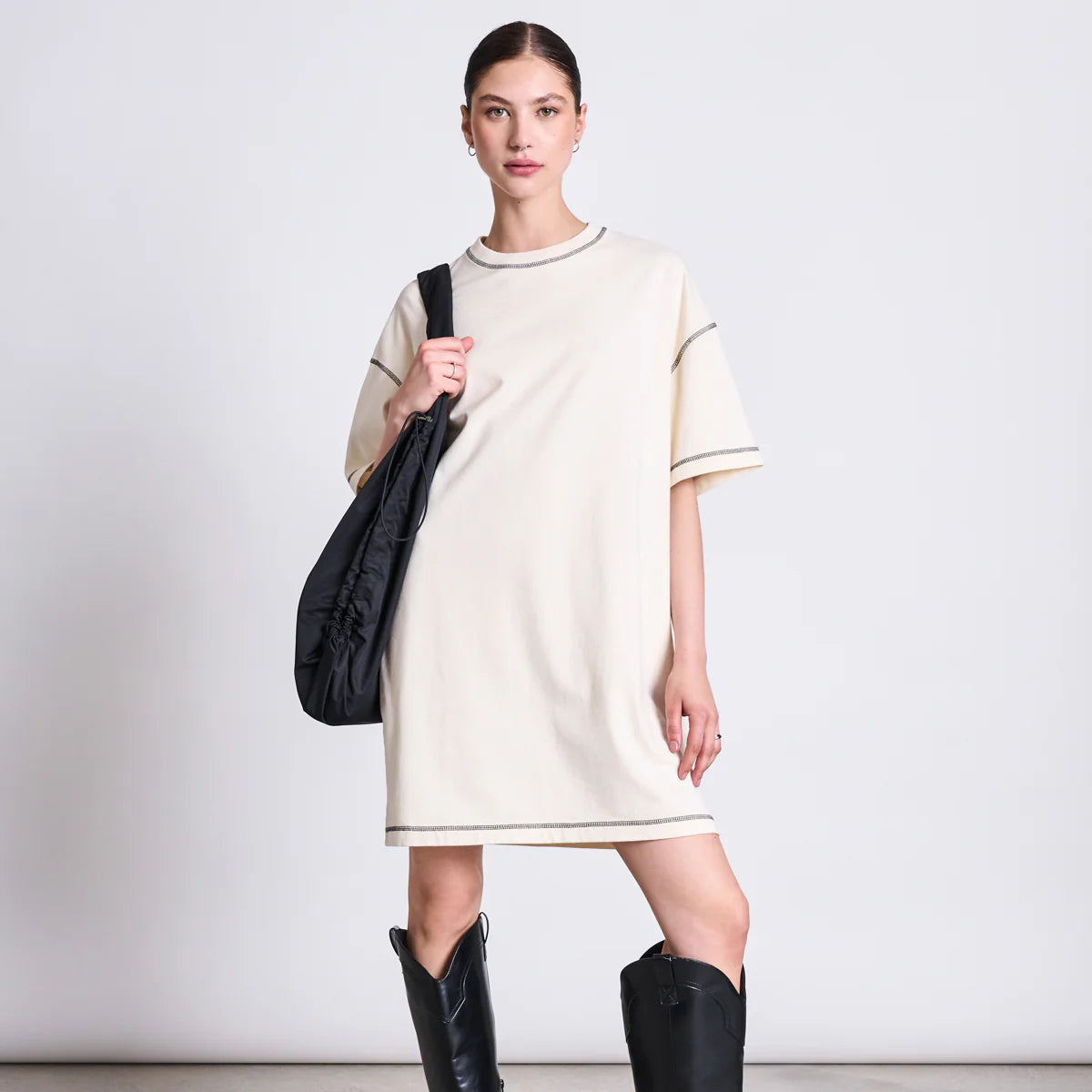 Eine Frau steht vor einem schlichten Hintergrund, trägt ein lockeres, kurzärmeliges, beigefarbenes T-Shirt-Kleid, schwarze, kniehohe Cowboystiefel und hat eine schwarze Umhängetasche dabei. Sie hat ihr Haar zurückgebunden und posiert mit einer Hand in der Hüfte.