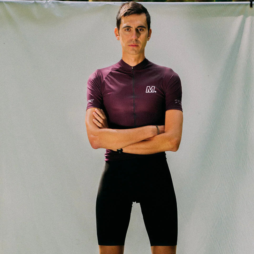 Eine Person in einem kastanienbraunen Fahrradtrikot und schwarzen Shorts steht mit verschränkten Armen vor einem hellen Hintergrund, in dem Bäume zu sehen sind.