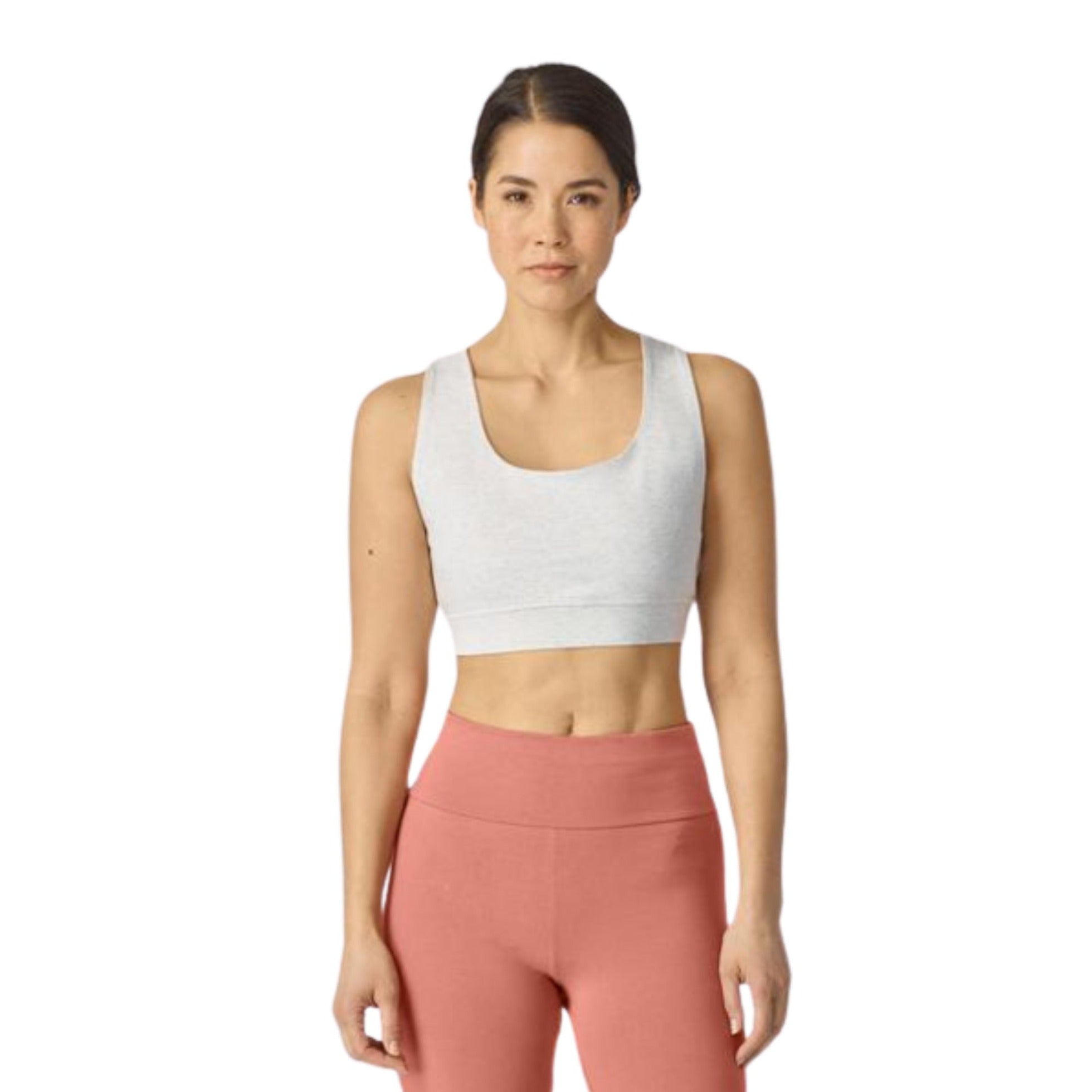 Frau in sportlichem Outfit: hellgraues Sportoberteil und rosafarbene Leggings, steht vor neutralem Hintergrund.
