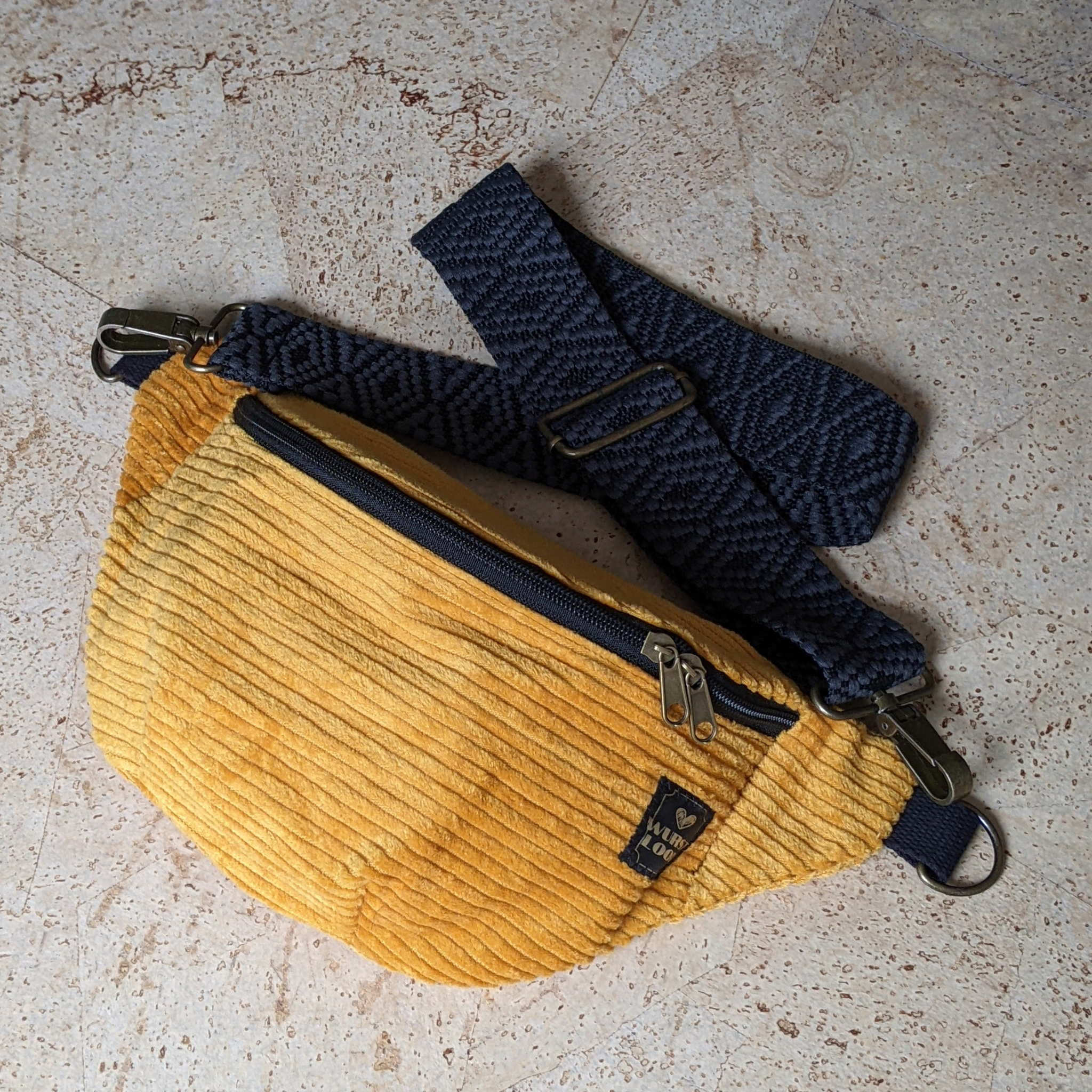 Eine gelbe Cord-Gürteltasche mit schwarzem Reißverschluss und einem dunkelblau gemusterten, verstellbaren Riemen liegt auf einer hellen Steinfläche.