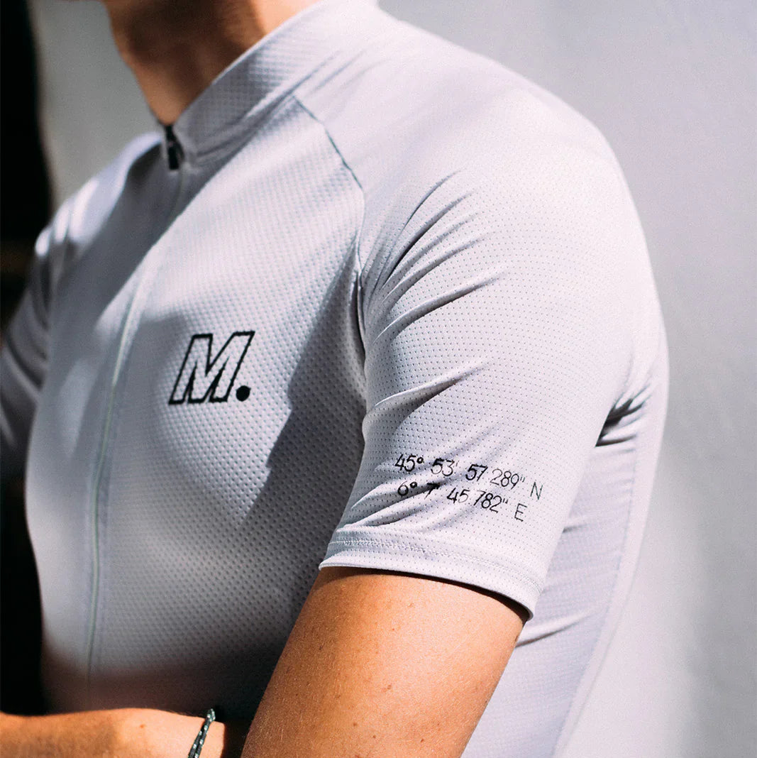 Eine Person trägt ein hellgraues, kurzärmeliges Radtrikot mit einem Reißverschluss, dem Aufdruck M. auf der Brust und geografischen Koordinaten auf dem Ärmel. Das Foto konzentriert sich auf den Torso und den entblößten Arm.