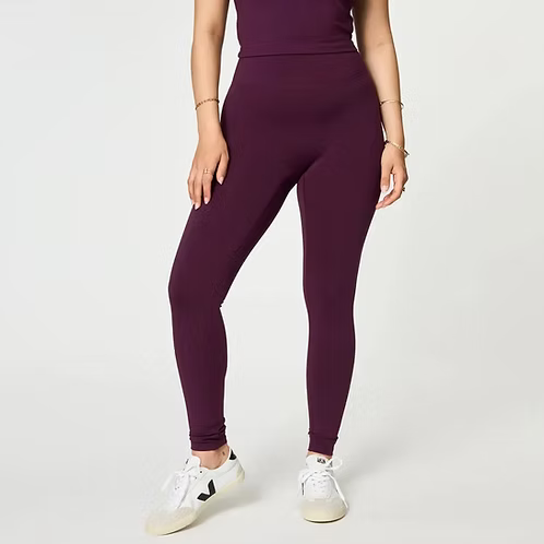 Eine Person, die ein passendes dunkelviolettes ärmelloses Oberteil und hochtaillierte Leggings sowie weiße Turnschuhe mit einem schwarzen V-Logo trägt, steht vor einem schlichten hellen Hintergrund. Nur der Körper von den Schultern abwärts ist sichtbar.