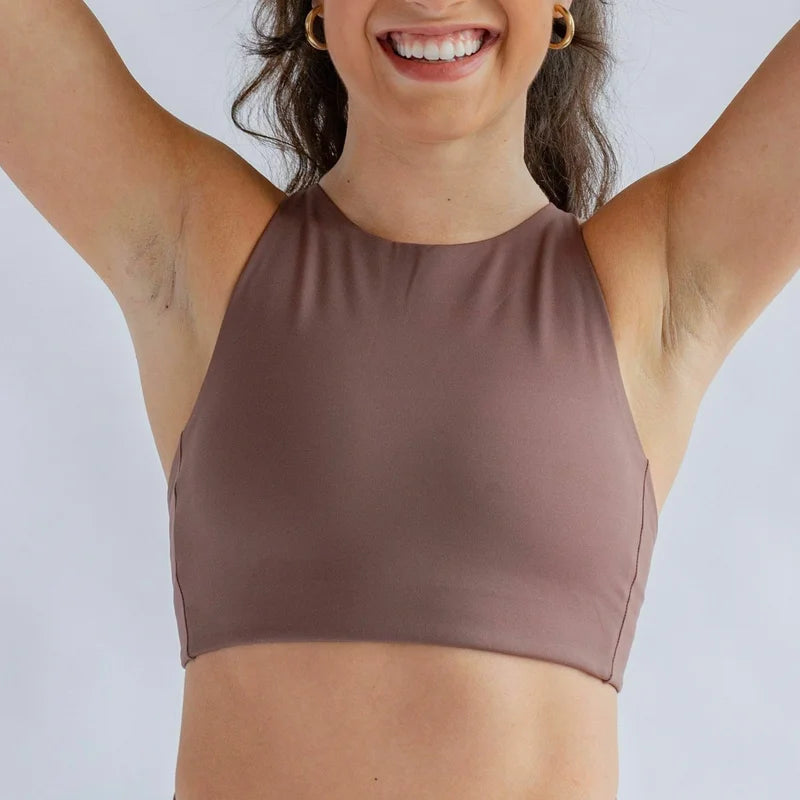 Eine Person in einem braunen ärmellosen Sportoberteil und dazu passenden hoch taillierten Leggings hebt die Arme und lächelt, während sie vor einem hellen Hintergrund steht.