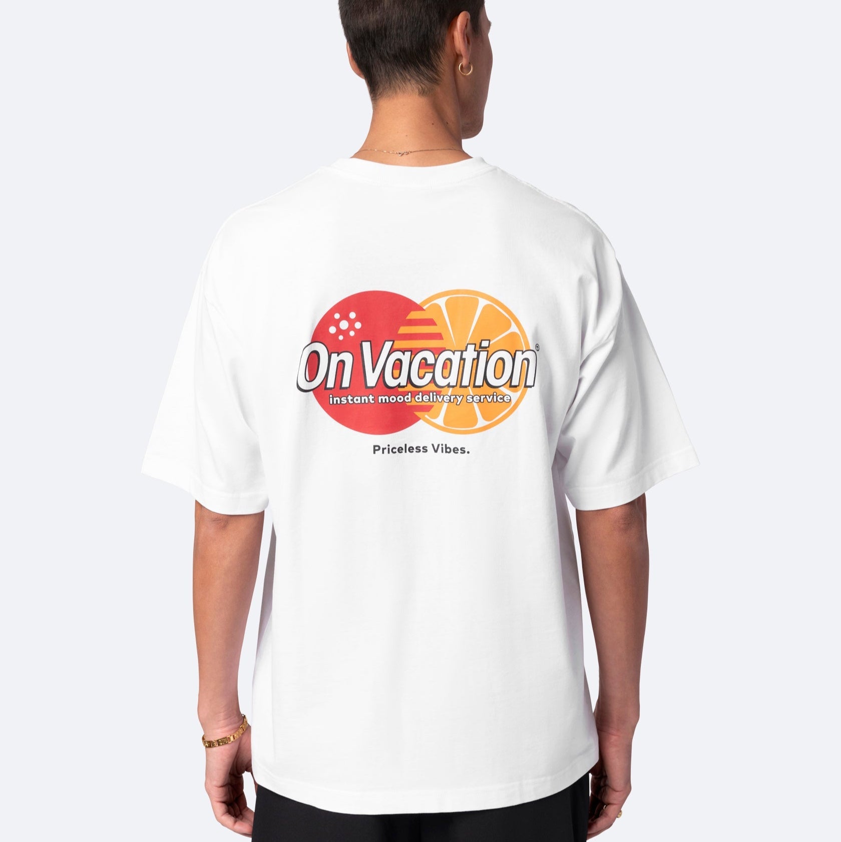 Eine Person steht mit dem Gesicht zur Wand und trägt ein weißes T-Shirt mit der Aufschrift "On Vacation" über orangefarbenen und roten Kreisen und der Aufschrift "Instant mood: daily travels and Priceless Vibes." darunter.
