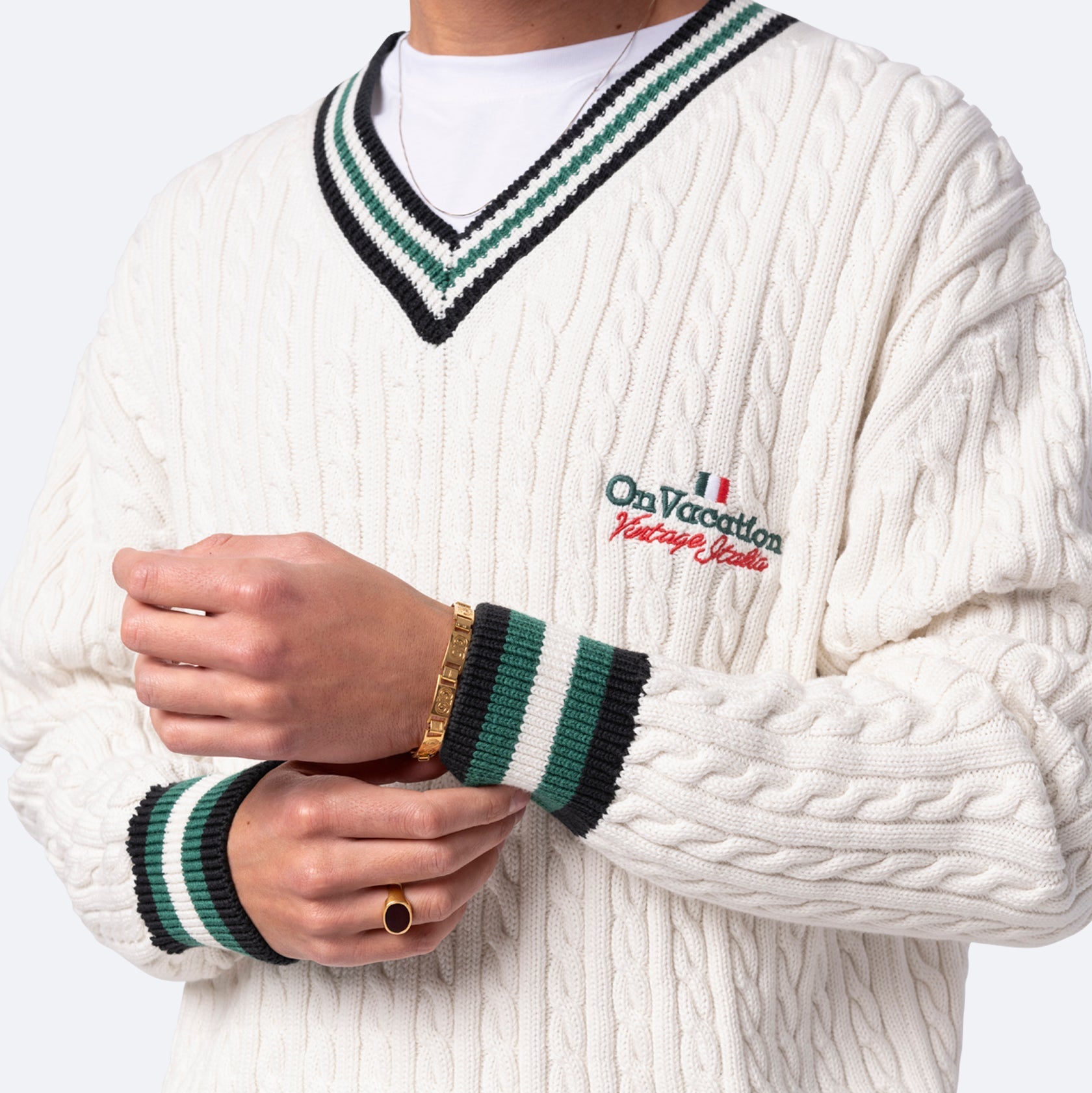 Eine Person, die einen weißen Zopfstrickpullover mit grün-schwarz gestreiften Manschetten und Kragen, einen gestickten Text mit der Aufschrift "On Vacation", ein goldenes Armband und einen schwarzen Ring trägt. Nur der Oberkörper und die Hände sind sichtbar.