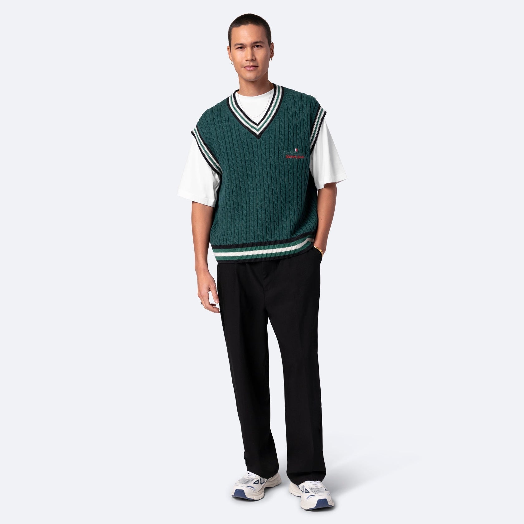 Ein Mann steht mit dem Gesicht nach vorne und trägt eine grüne Strickweste über einem weißen kurzärmeligen Hemd, eine schwarze Hose und weiße Turnschuhe vor einem schlichten hellgrauen Hintergrund.