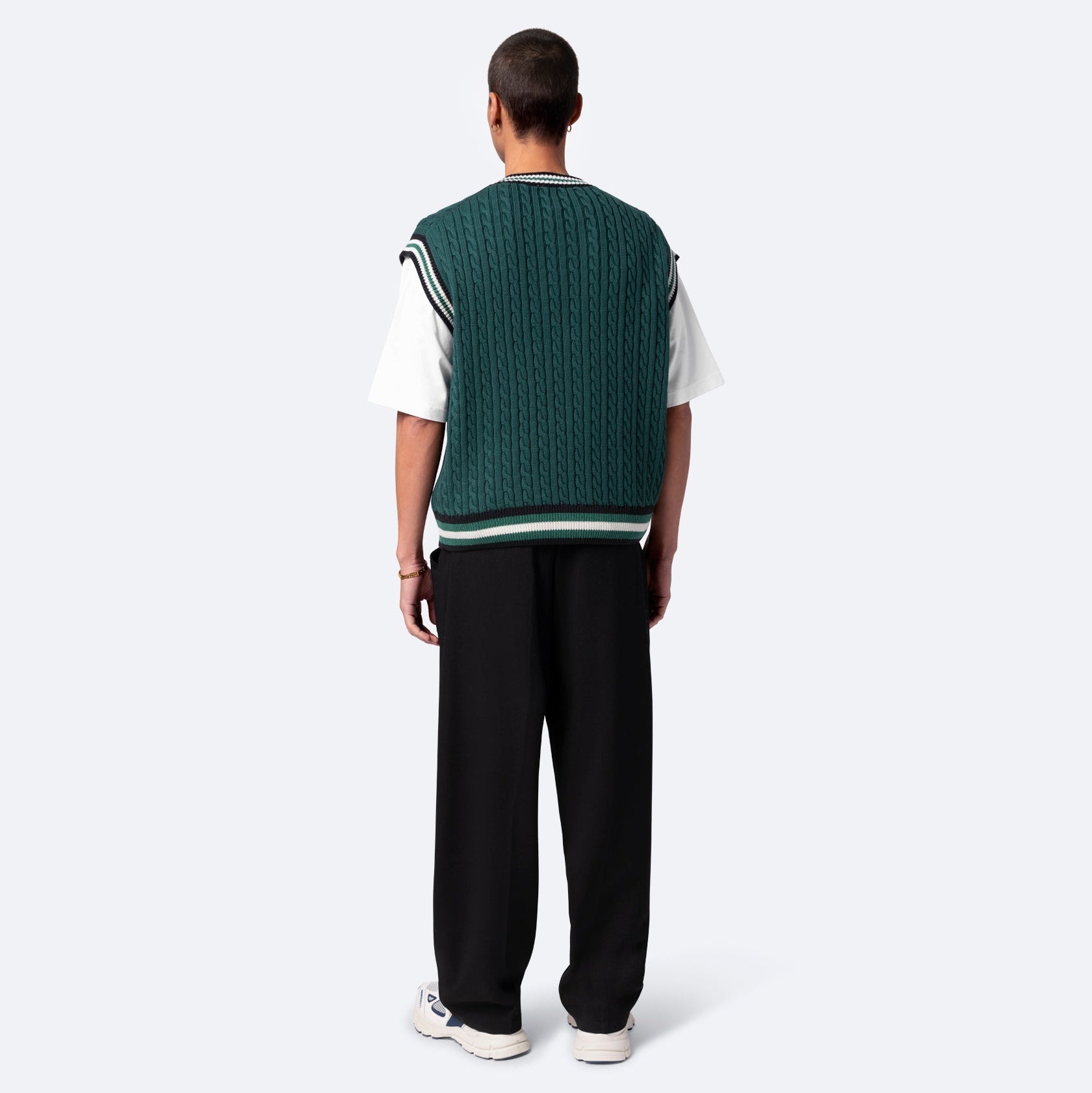 Eine Person steht mit dem Gesicht nach hinten, trägt eine grüne Strickweste über einem weißen T-Shirt, eine weite schwarze Hose und weiße Turnschuhe vor einem schlichten hellen Hintergrund.