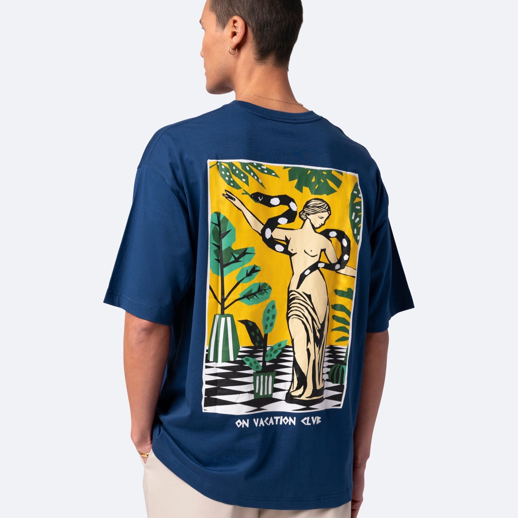 Eine Person trägt ein blaues T-Shirt mit einer farbenfrohen Grafik einer klassischen Statue, Pflanzen und einer Schlange auf dem Rücken, zusammen mit dem Text ON VACATION CLUB am unteren Rand des Designs.