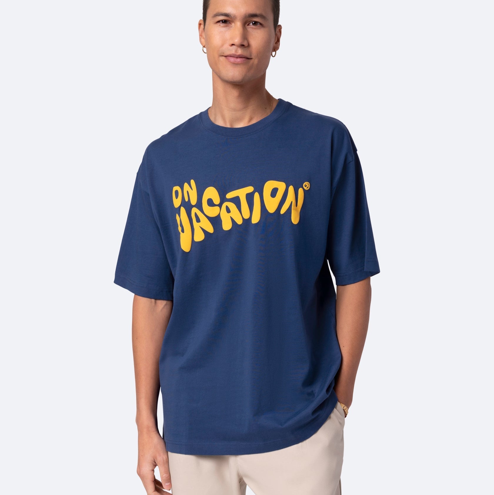 Eine Person, die ein marineblaues T-Shirt mit der Aufschrift ON VACATION in großen, gewellten gelben Buchstaben trägt, gepaart mit einer hellbeigen Hose, steht vor einem schlichten hellen Hintergrund.