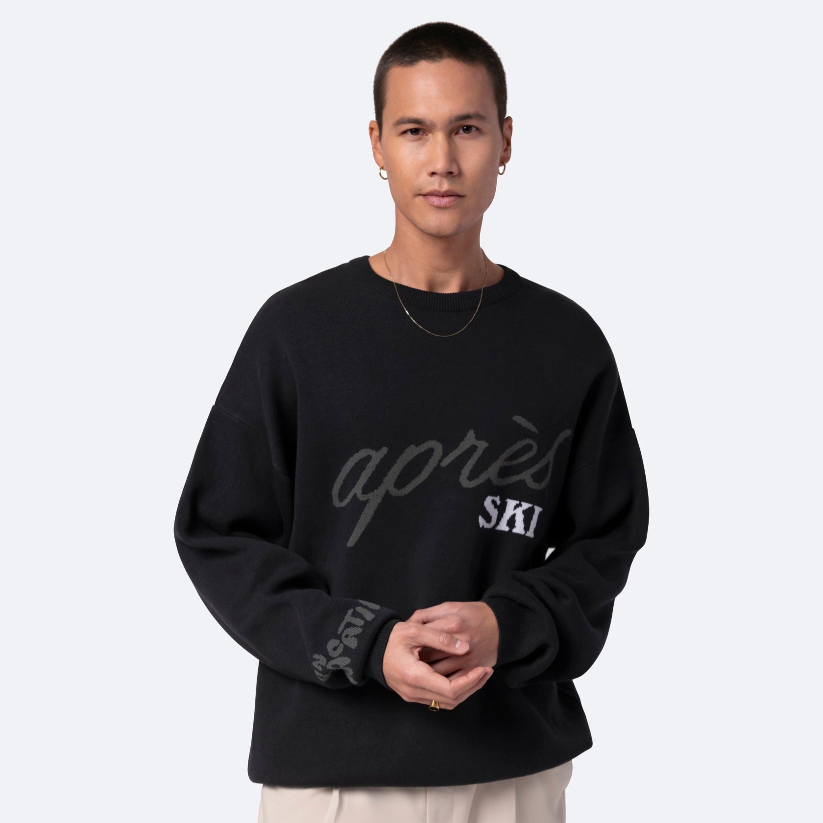 Eine Person mit kurzen Haaren, die ein schwarzes Sweatshirt mit der Aufschrift après SKI und eine cremefarbene Hose trägt, steht vor einem hellgrauen Hintergrund.