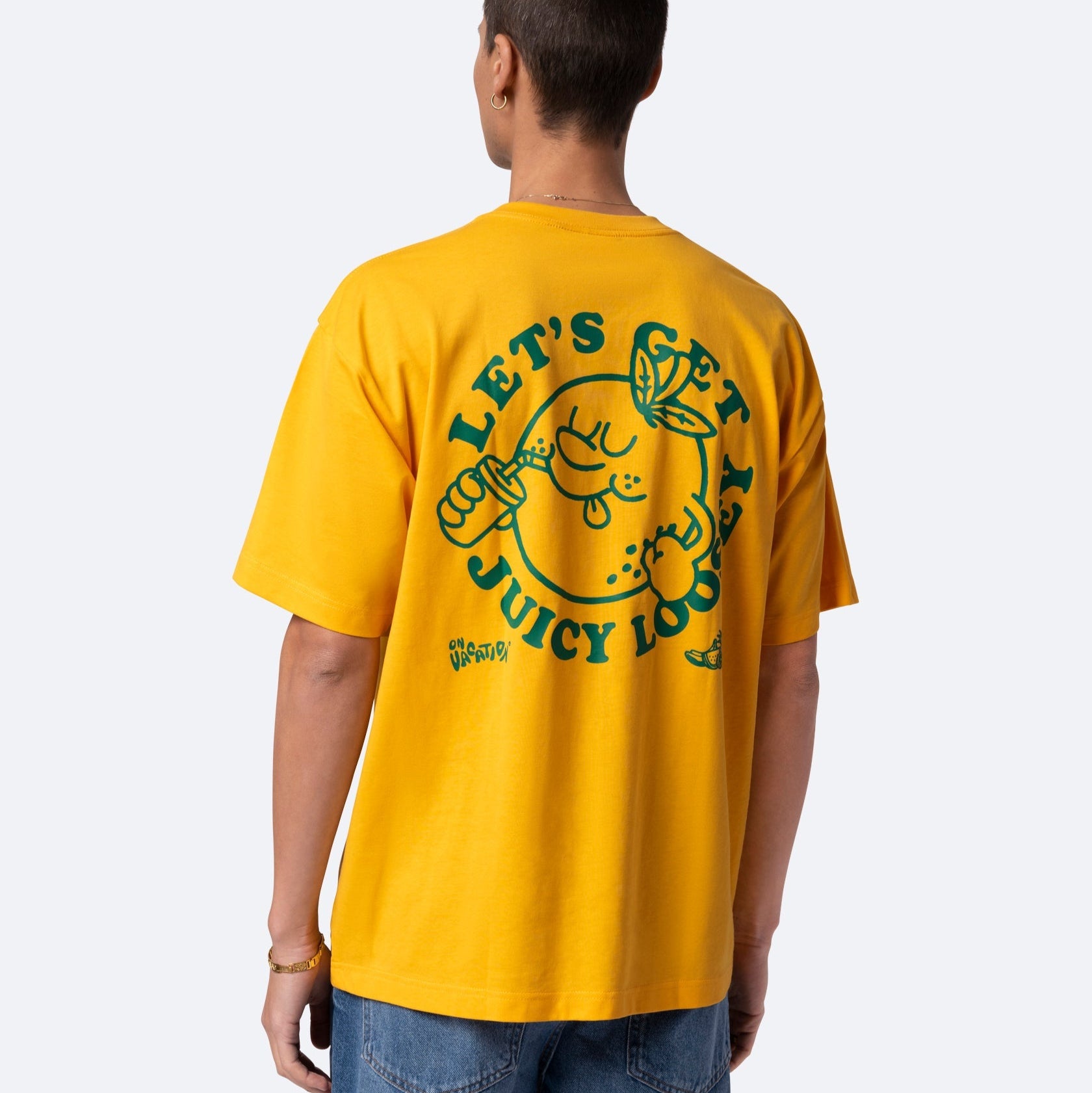 Eine Person trägt ein gelbes T-Shirt mit einer Cartoon-Zitrone, die ein Getränk hält, und der Aufschrift LETS GET JUICY LOOT in grüner Schrift auf dem Rücken. Die Person blickt weg und trägt eine blaue Jeans.