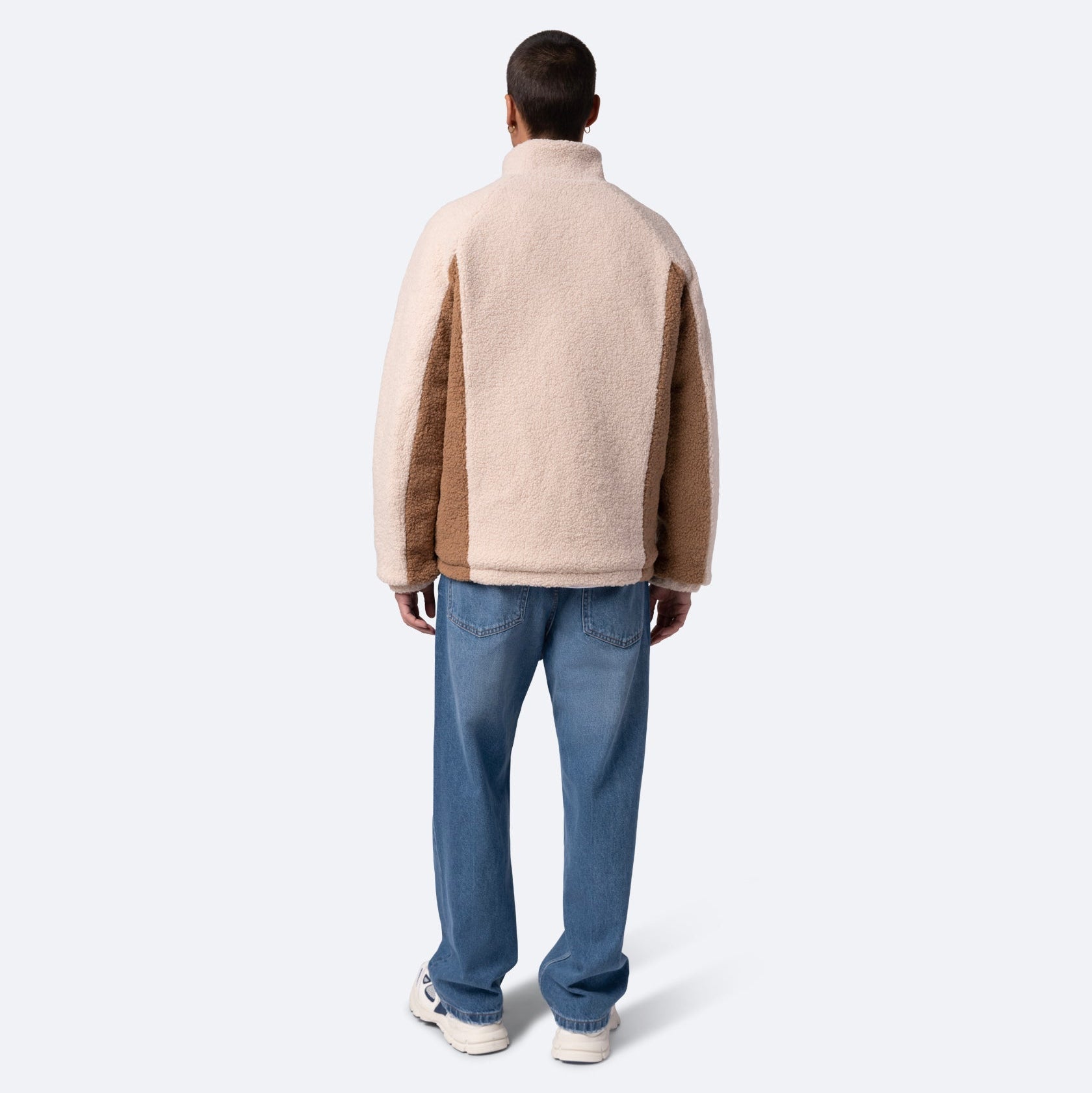 Eine Person steht mit dem Gesicht zur Wand, trägt eine beige-braune Fleecejacke, blaue Jeans und weiße Turnschuhe vor einem schlichten hellen Hintergrund.