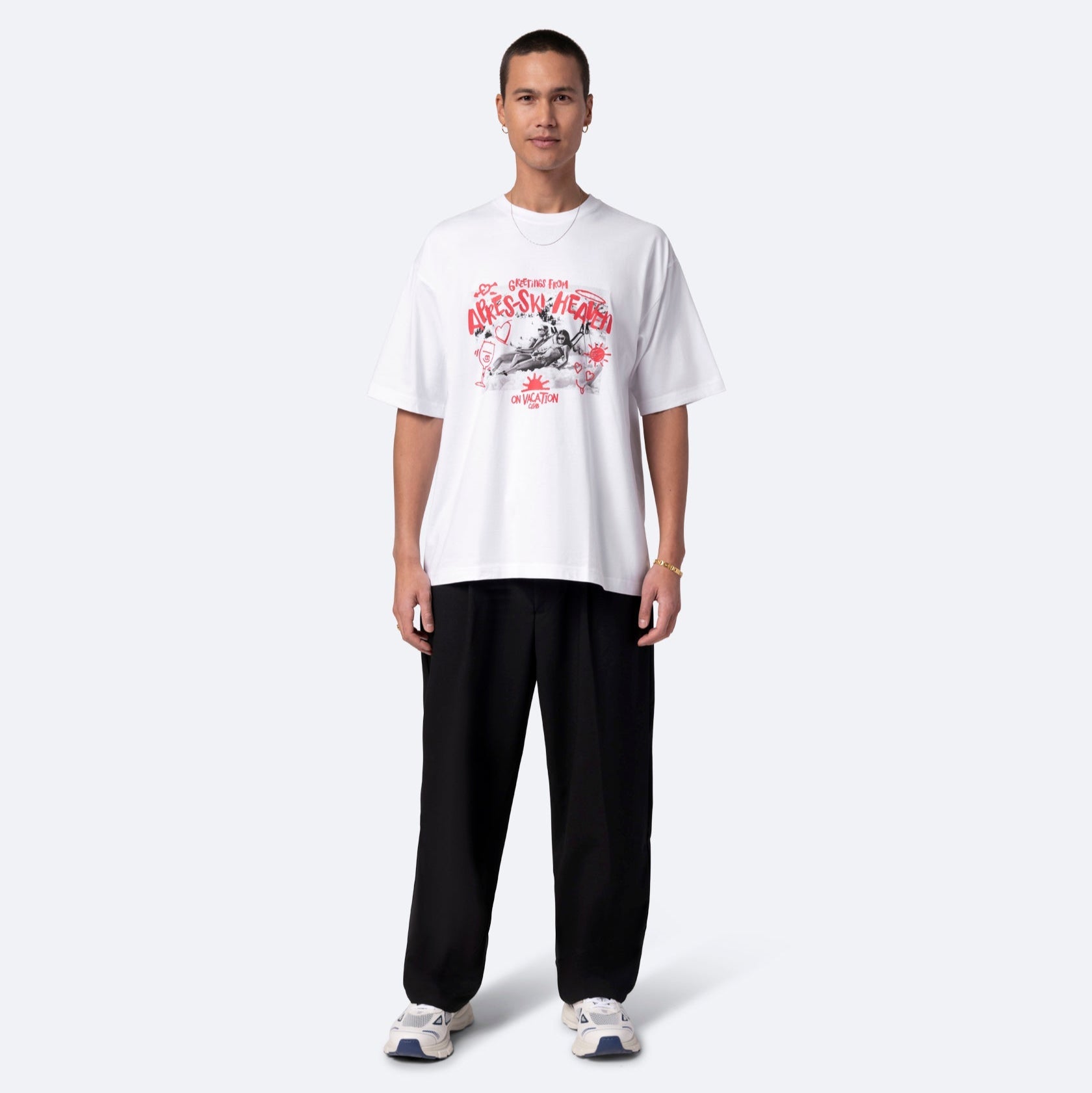 Eine Person steht mit dem Gesicht nach vorne und trägt ein lockeres weißes T-Shirt mit roten und schwarzen Motiven, eine schwarze Hose und weiße Turnschuhe vor einem schlichten hellgrauen Hintergrund.