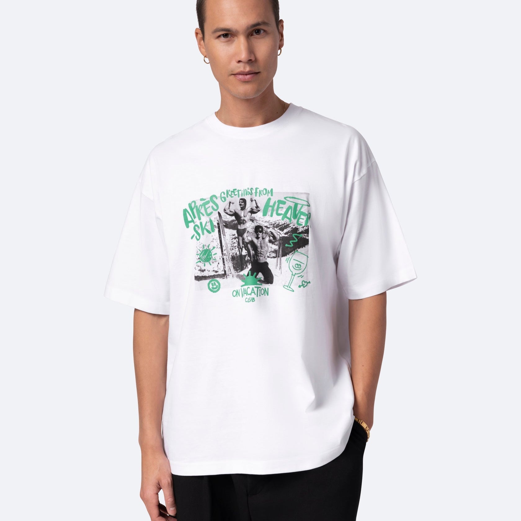 Ein Mann mit kurzen Haaren, der ein lockeres weißes T-Shirt mit grünem und schwarzem Text und Bildern trägt, steht mit einer Hand in der Tasche vor einem schlichten hellen Hintergrund.