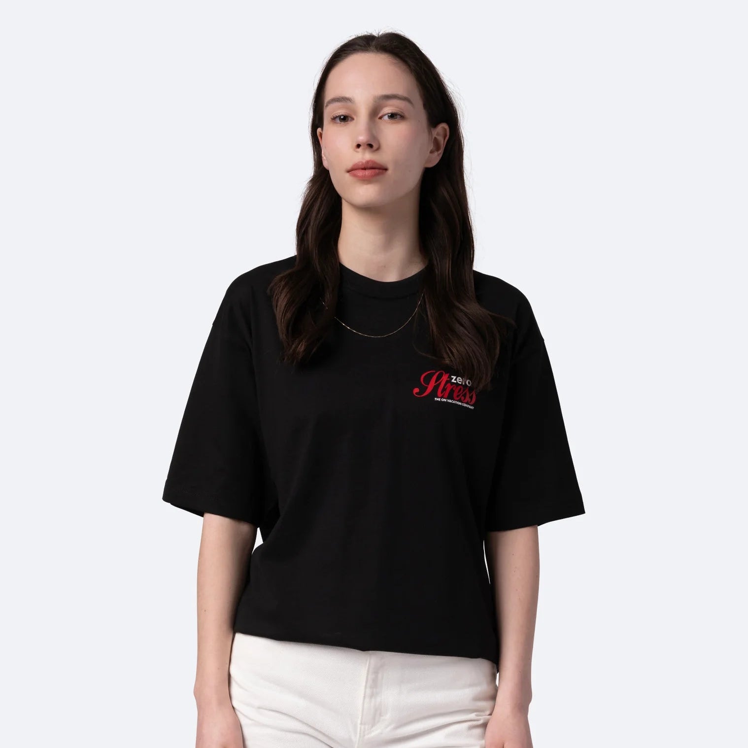 Eine Frau mit langen braunen Haaren trägt ein schwarzes übergroßes T-Shirt mit roter Aufschrift auf der Brust und eine weiße Hose vor einem schlichten hellgrauen Hintergrund.
