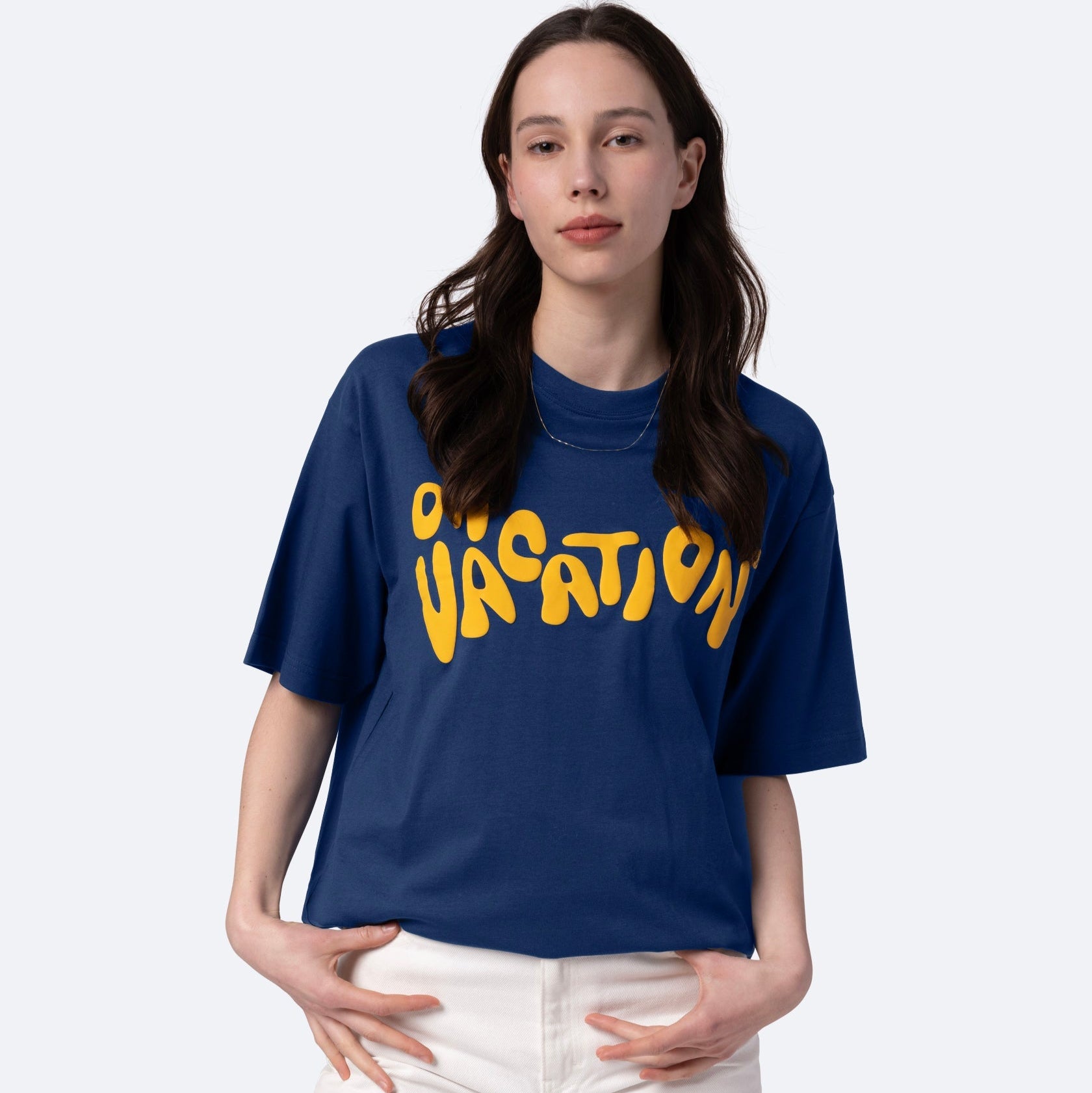 Eine Frau mit langen braunen Haaren trägt ein blaues T-Shirt mit dem Wort VACATION in fetten gelben Buchstaben und eine weiße Hose. Sie steht vor einem schlichten hellen Hintergrund und schaut mit neutralem Blick in die Kamera.
