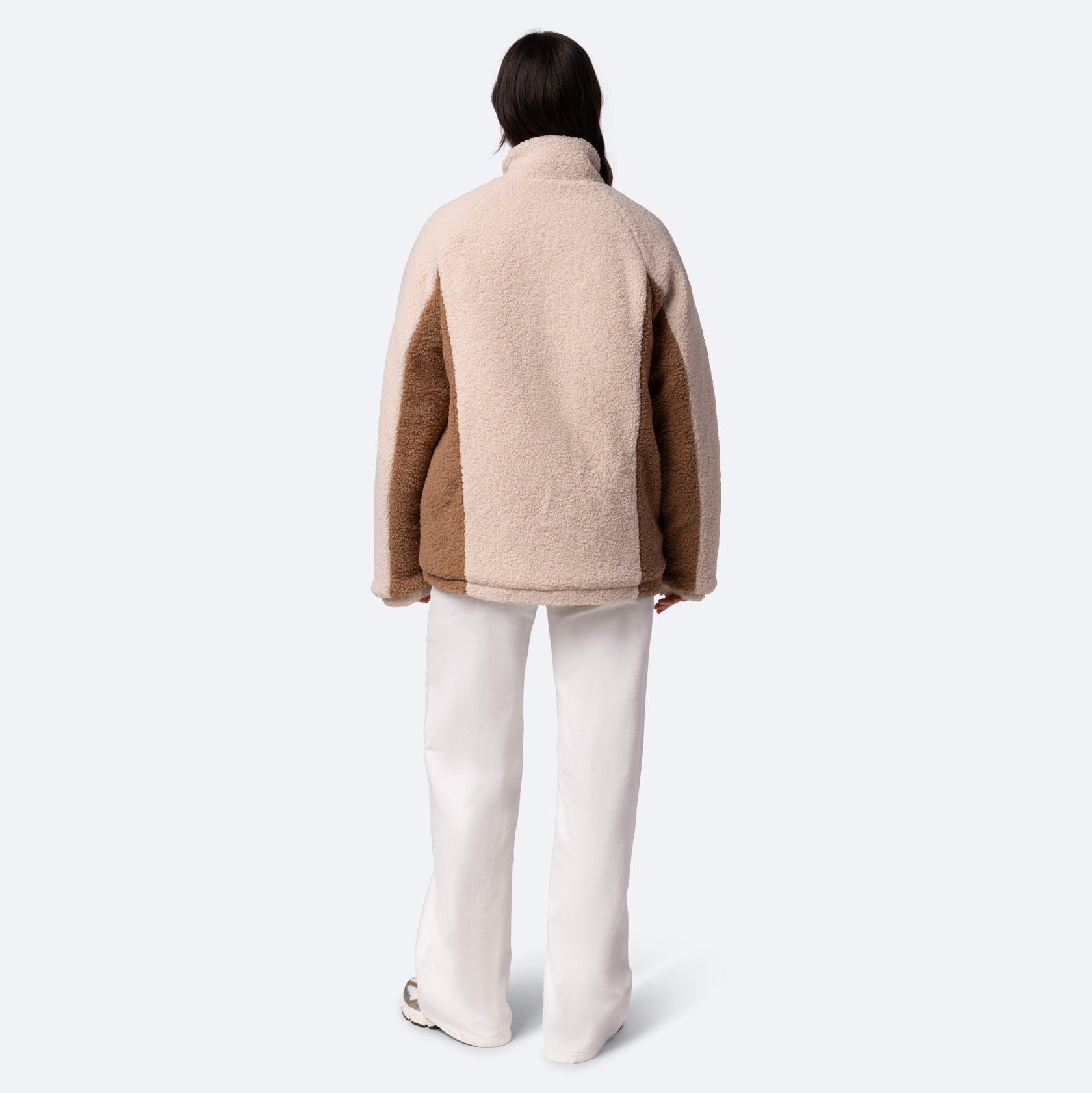 Eine Person mit langen dunklen Haaren trägt eine beige-braune Fleecejacke und eine weiße Hose und steht mit dem Rücken zur Kamera vor einem schlichten hellen Hintergrund.